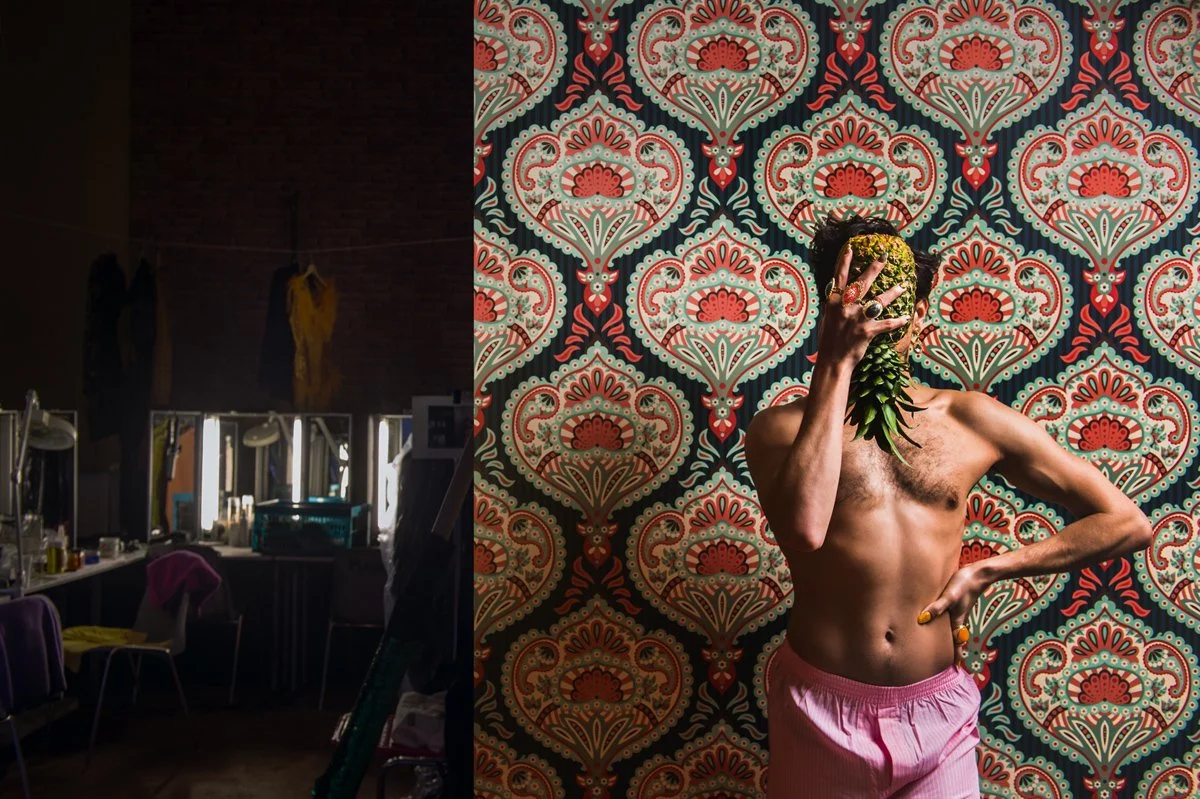 Ein Mensch posiert vor einer Wand mit blumigem Muster, hält eine Ananas vor das Gesicht und trägt pinke Shorts.