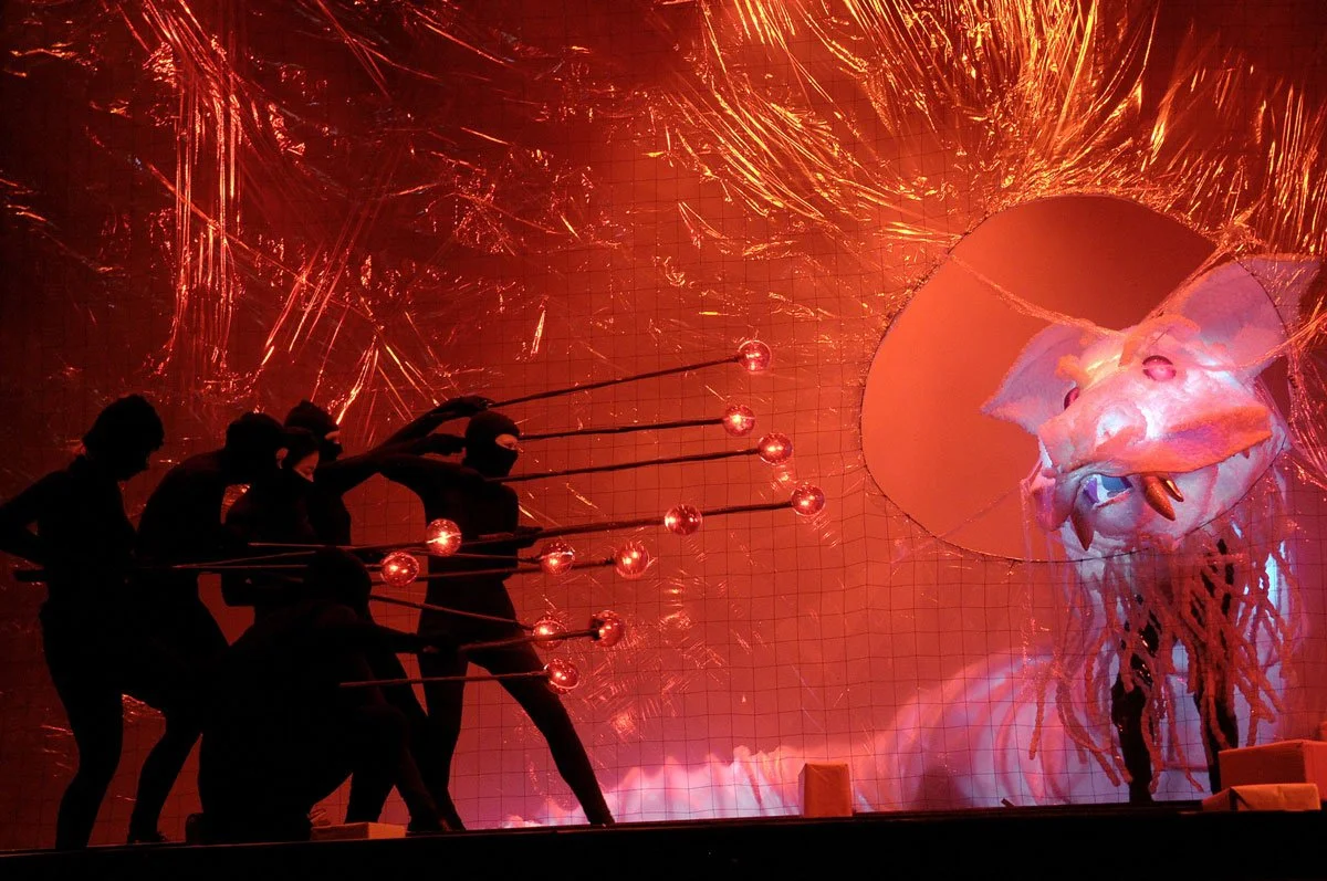 Schatten von fünf Menschen, die mit Lichtern spielen, vor einer leuchtenden roten Hintergrundkulisse mit einem großen Drachenkopf als Bühnendekoration.