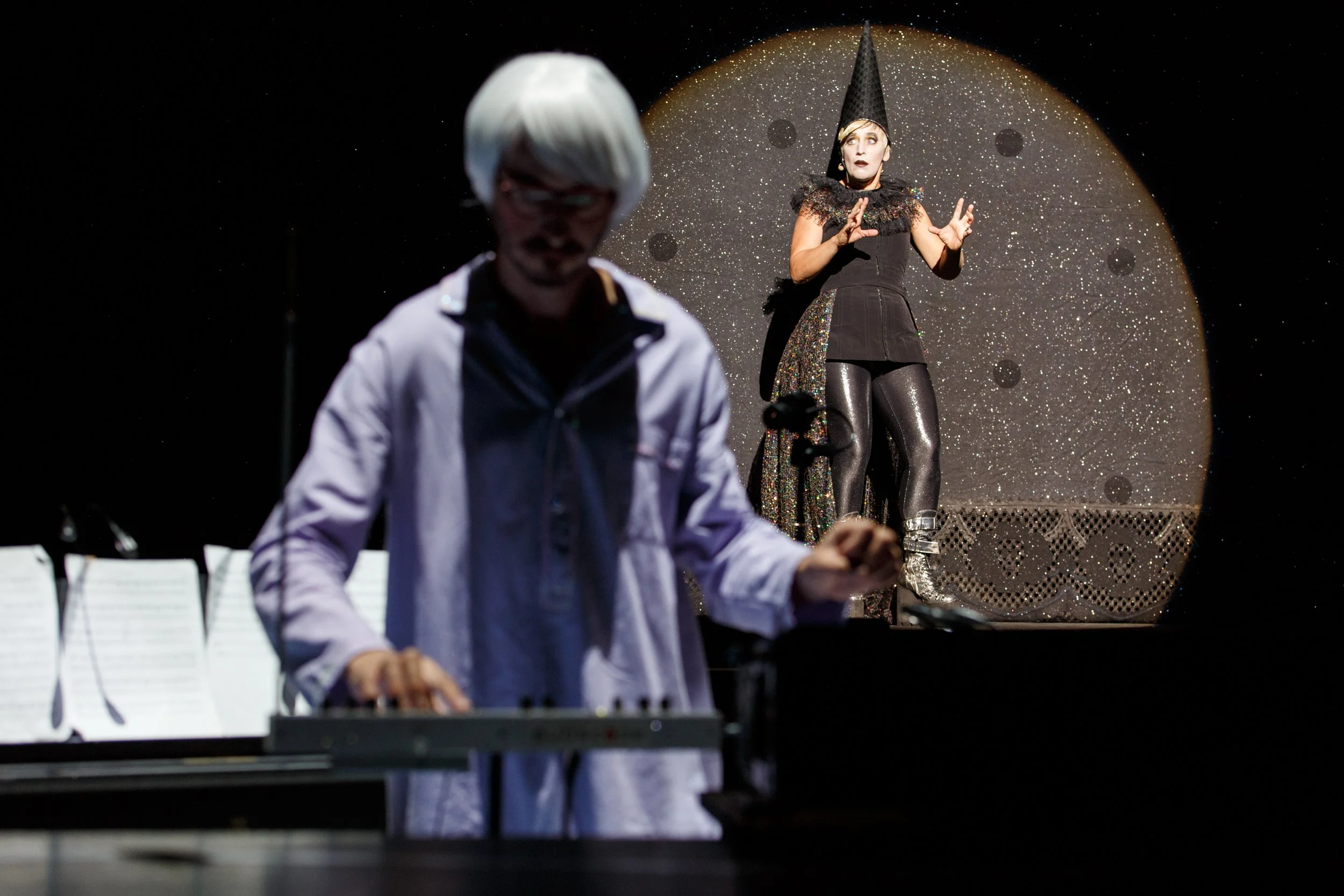 Bühnenaufnahme mit einem Pianisten im Vordergrund und einer performenden Frau im Hintergrund. Die Frau trägt ein schwarzes Outfit mit glitzernden Leggings und einem spitzen Hut, während der Pianist eine weiße Perücke trägt.