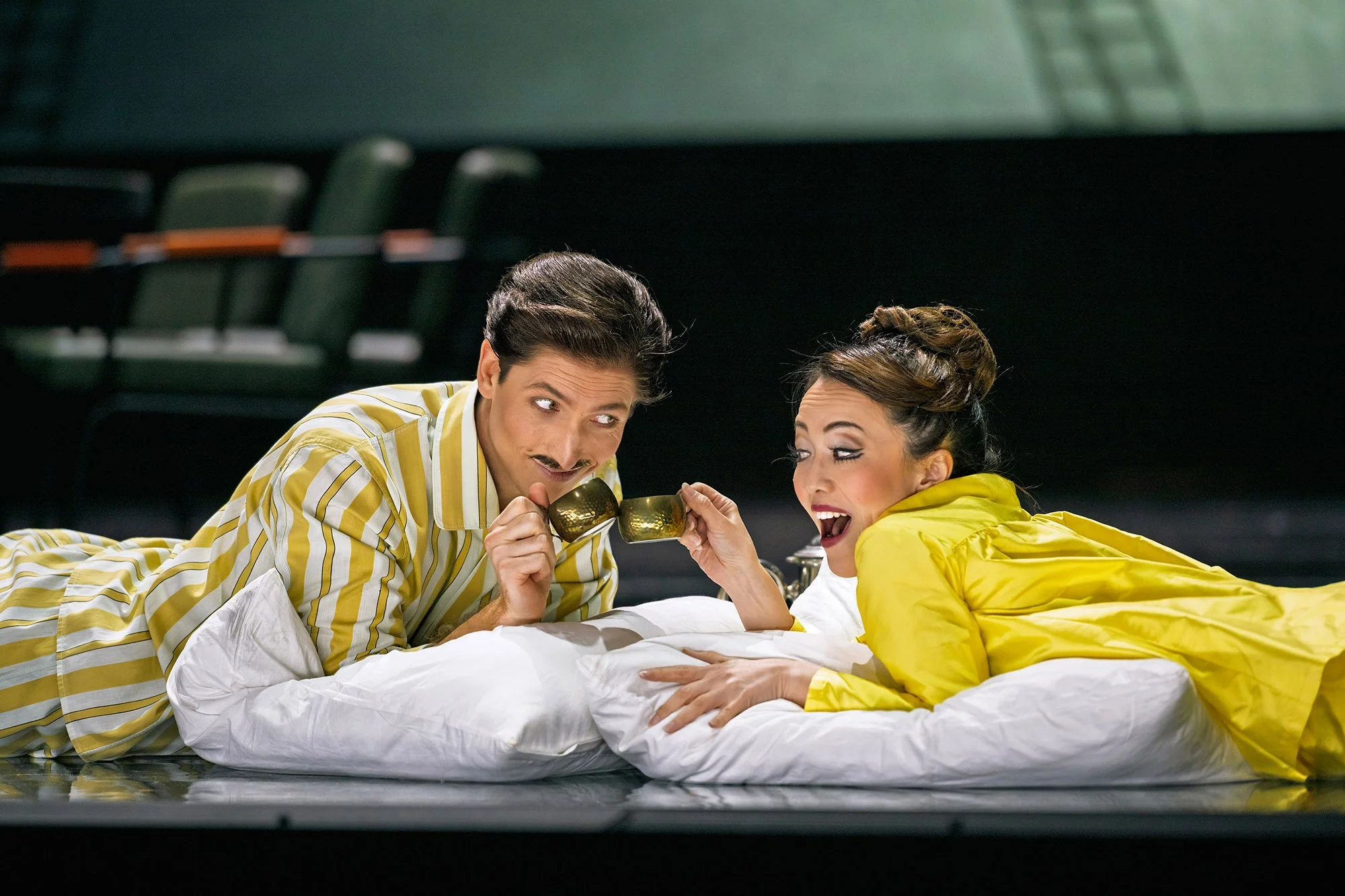 Zwei Menschen liegen auf einem Bett, trinken aus Goldtassen, lachen und scheinen eine Szene aus einem Theaterstück oder Film zu spielen, mit dunklem Hintergrund.