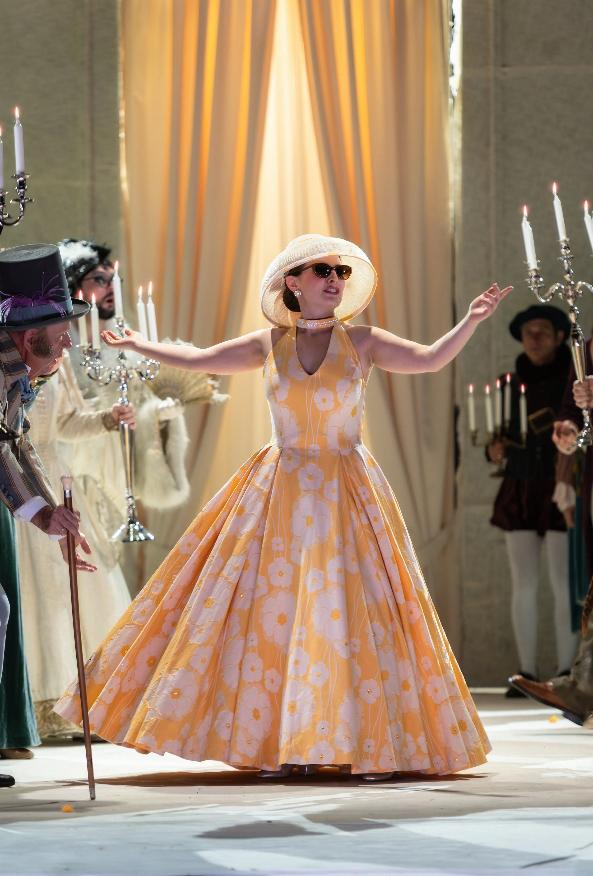 Eine Frau in einem langen, gelben, blumenmusternden Kleid mit großen Polizisten in der Mitte, die sich gegenseitig umarmen und tanzen, umgeben von anderen Personen in historischen Kostümen. Sie trägt eine große Sonnenblende und Sonnenbrille.