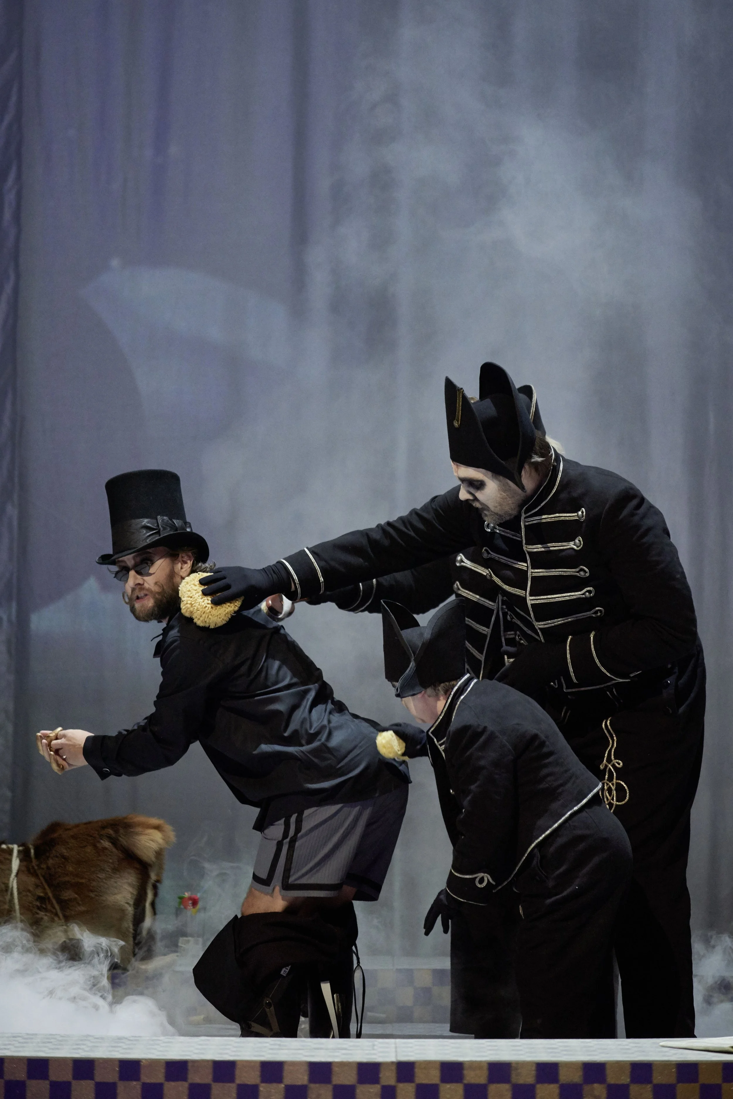 Drei Schauspieler in schwarzen Kostümen und Zylindern spielen eine Szene mit Rauch und einem Tier auf einer Bühne.