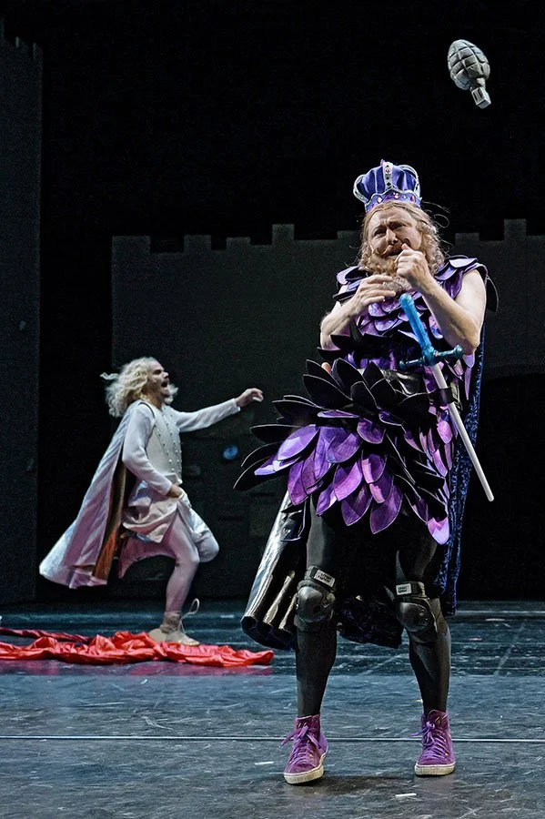 Zwei Schauspieler auf einer Bühne, einer trägt ein königliches Kostüm mit einem roten Bart, der andere trägt ein Fantasiekostüm mit Flügeln und Knieprotektoren, in einer Theaterszene mit dunklem Hintergrund.