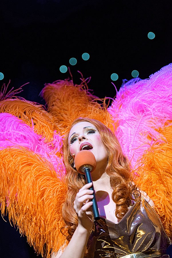 Eine Frau mit roten, gewellten Haaren singt in einem Mikrofon vor einem farbenfrohen Hintergrund mit großen, leuchtenden Pfauenfedern in Pink, Orange und Lila.
