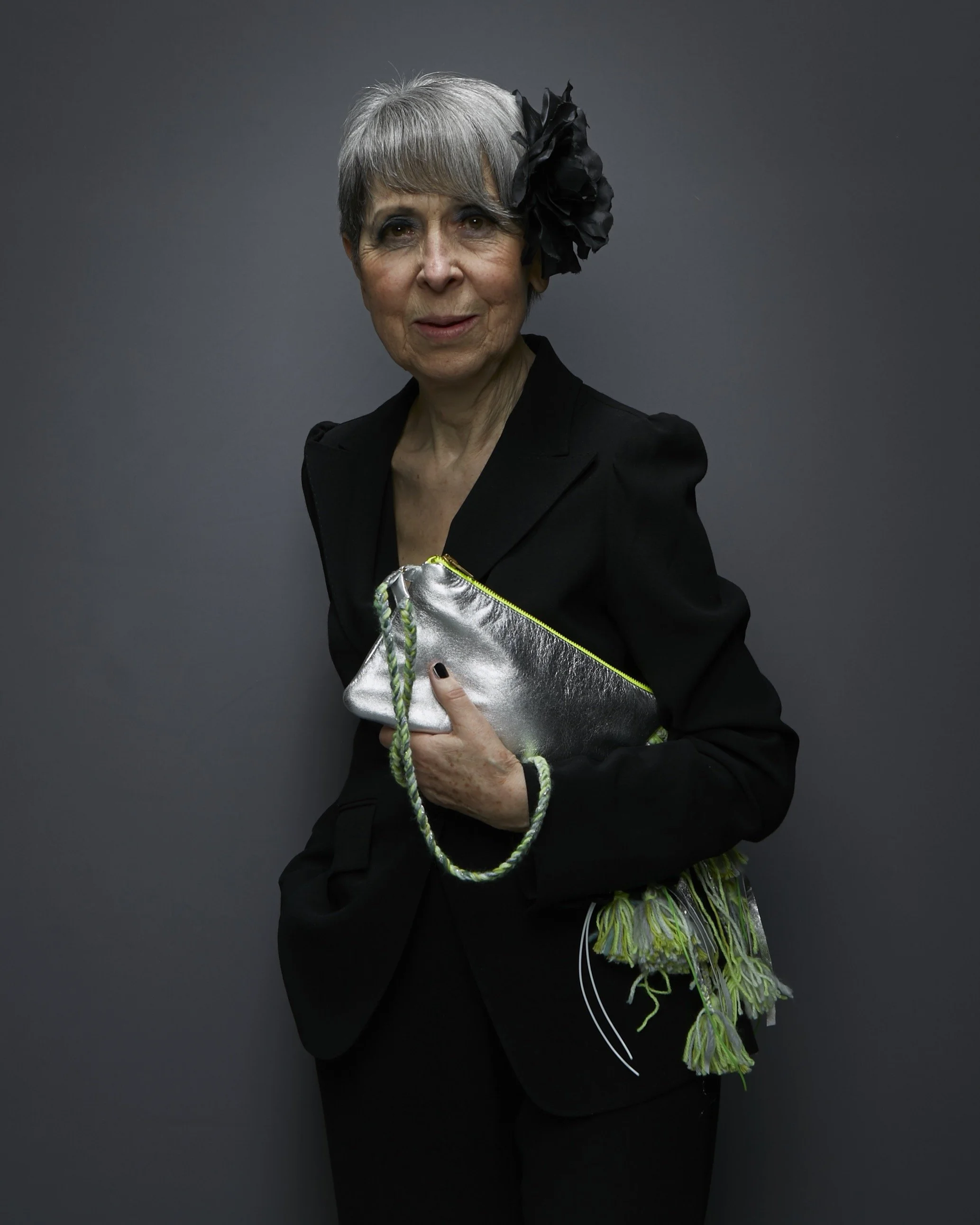 Eine ältere Frau mit kurzen grauen Haaren trägt schwarze Kleidung, hat eine große schwarze Blume im Haar und hält eine silberne Clutch. Sie steht vor einem dunklen grauen Hintergrund.