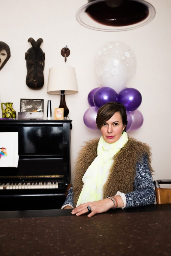 Eine Frau (Dinah Ehm) sitzt an einem Tisch vor einem Klavier mit einer bunten Zeichnung auf dem Notenblatt. Im Hintergrund befinden sich Dekorationen, einschließlich Ballons, einer Lampe, einer Masken und einer Stofffigur.