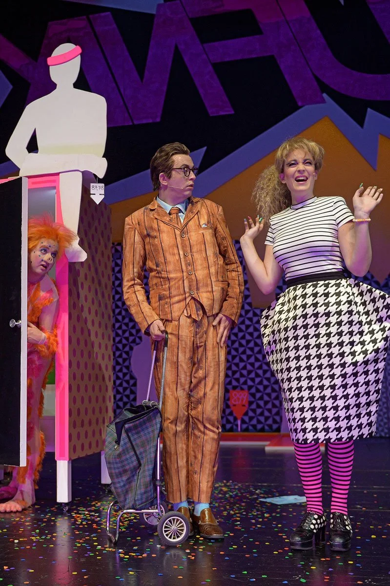 Schauspieler in farbenfroher Kostümierung auf der Bühne während eines Theaterspiels, mit einer bunten Kulisse und Konfetti auf dem Boden.