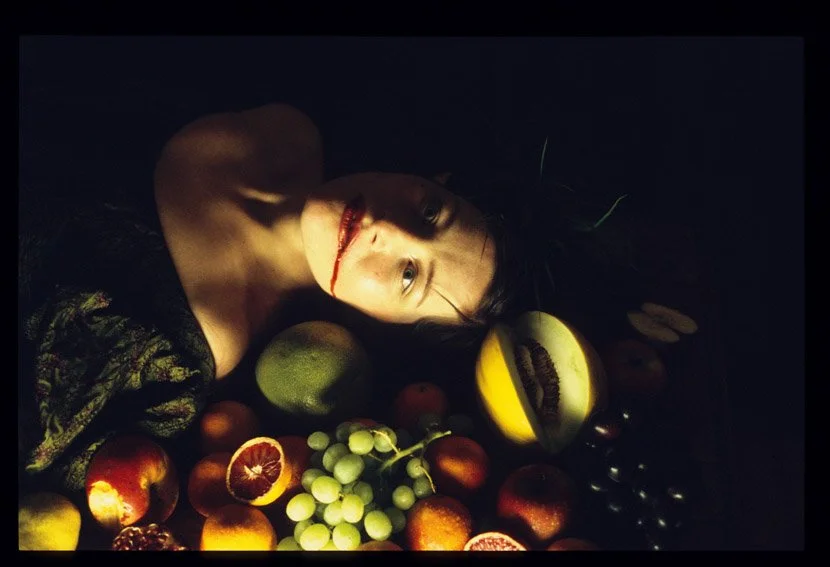 Eine Person liegt auf einem Tisch voller verschiedenartiger Früchte. Die Person hat einen Schnitt im Mund, der blutet, und schaut nach oben. Die Umgebung ist dunkel.