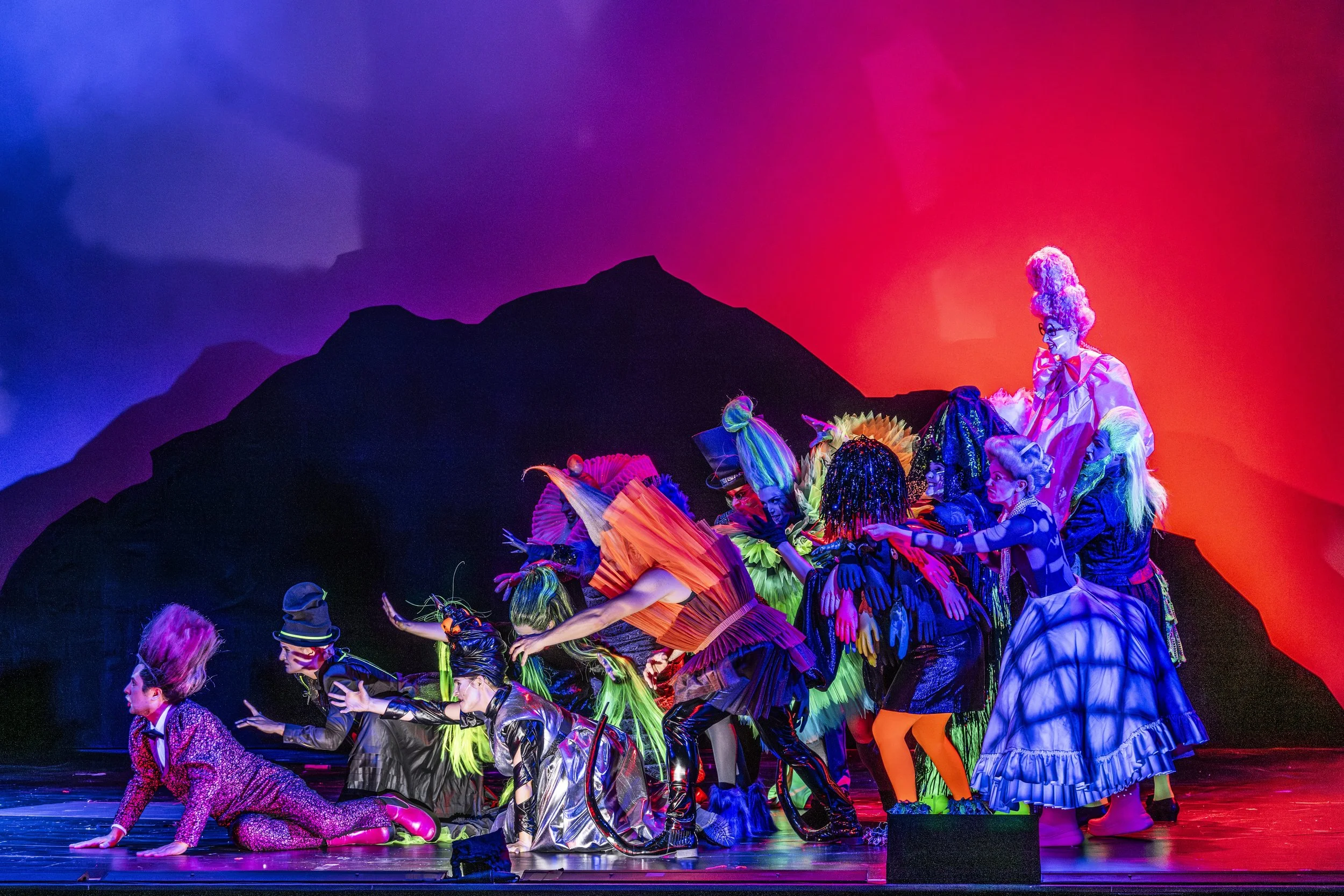 Eine bunte Theaterbühne mit zahlreichen Darstellern in fantasievollen Kostümen, die eine Szene aus einem Theaterstück oder Musical darstellen, vor einem dunklen Berg im Hintergrund mit rotem und blauen Licht