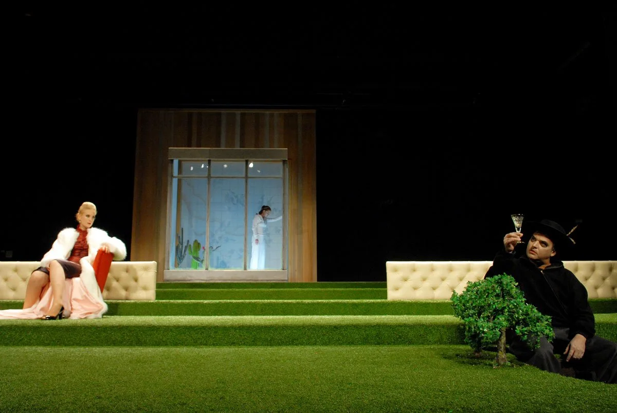 Theaterbühne mit drei Schauspielern: eine Frau sitzt auf einer Couch links, eine Person steht vor einem Fenster in der Mitte, und ein Mann sitzt rechts mit einem Glas in der Hand, neben einem Busch.