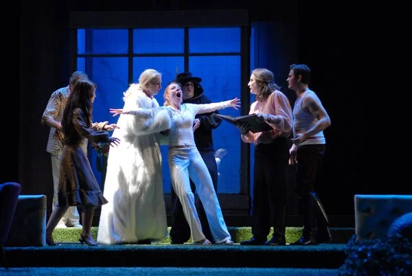 Schauspieler auf einer Bühne, die eine dramatische Szene mit mehreren Personen darstellt, vor einem großen Fenster mit blauer Beleuchtung im Hintergrund.