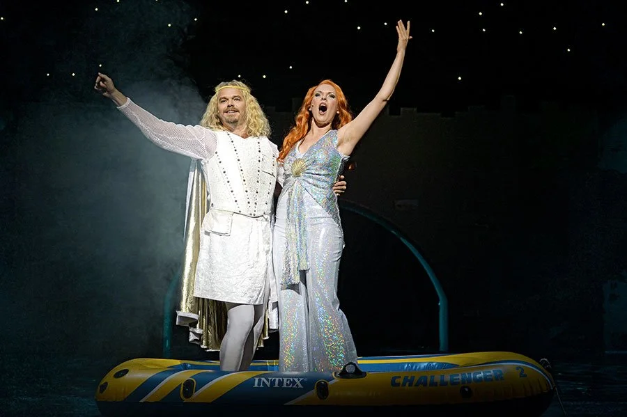 Zwei Menschen in bunten Kostümen auf einer Bühne, ein Mann in weißem Gewand mit langem blonde Haar und eine Frau in einem schimmernden Kleid, stehen auf einem gelben Wasserfahrzeug, vermutlich in einer Theater- oder Show-Performance.