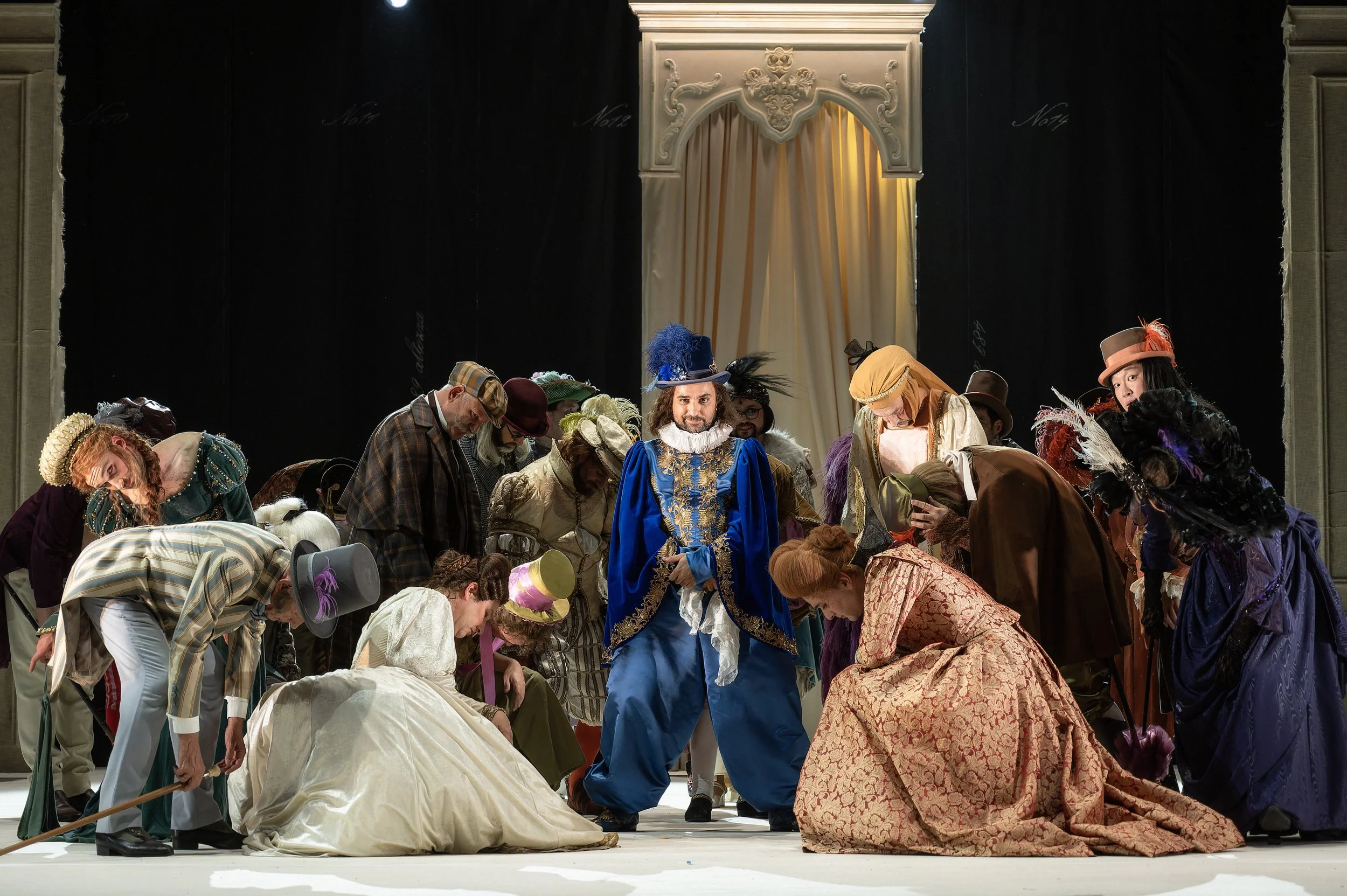 Bühnenbild mit Menschen in historischer Kleidung bei einer Theateraufführung, zentrale Figur trägt ein blaues Kostüm mit goldenen Verzierungen, im Hintergrund eine Palastarchitektur