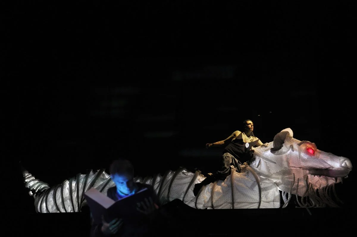 Zwei Kinder auf einem fantastischen Drachen- und Meereswesen-Kostüm bei einer Theateraufführung, dunkler Hintergrund, eine Kerze auf dem Kopf des Wesens. Das Wesen hat leuchtende rote Augen.