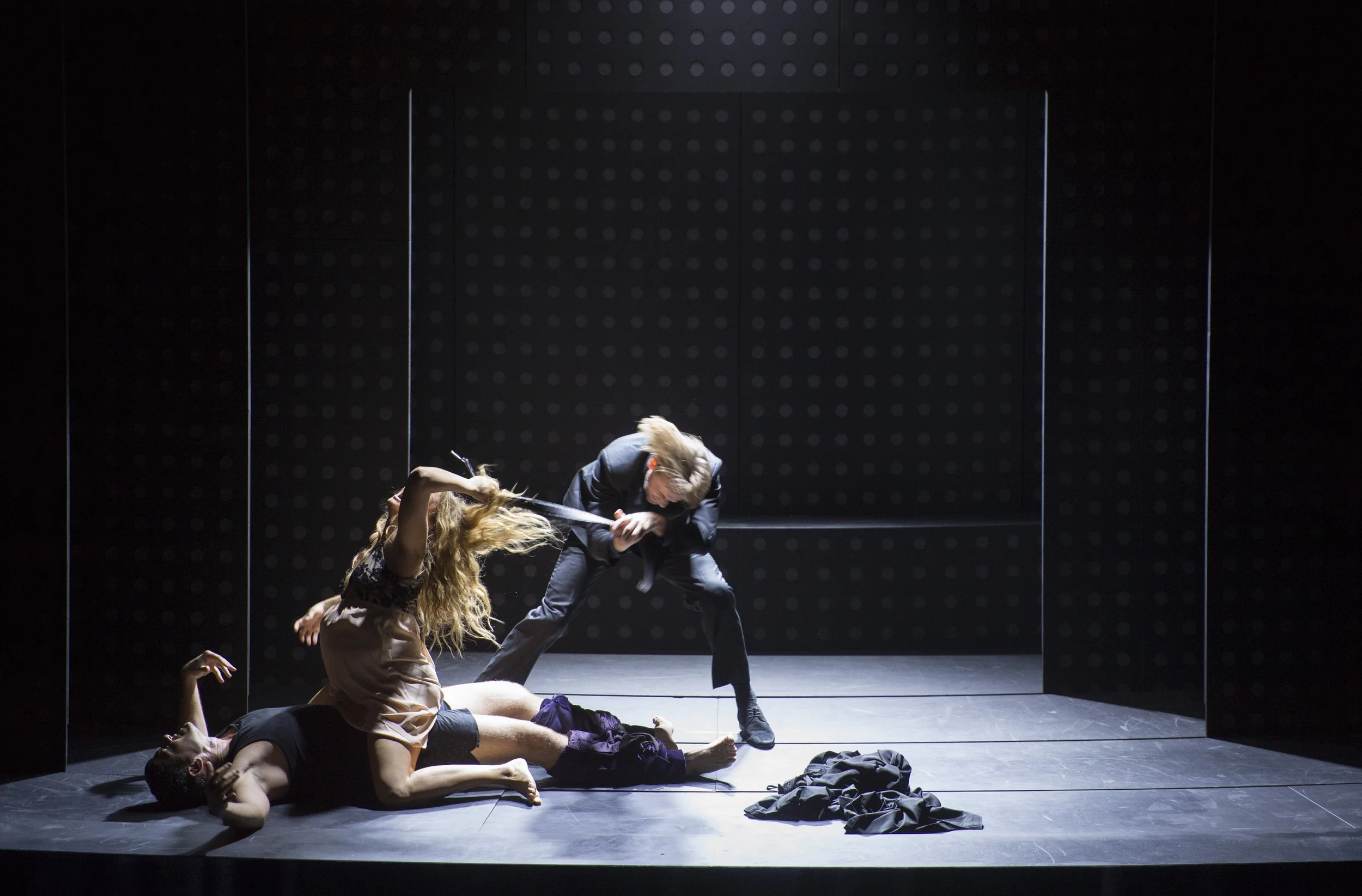 Drei Menschen auf einer Bühnenfläche, eine Person liegt auf dem Boden, eine andere ist oben auf dieser Person, während die dritte Person mit einem Schwert auf die Person auf dem Boden einschlägt.