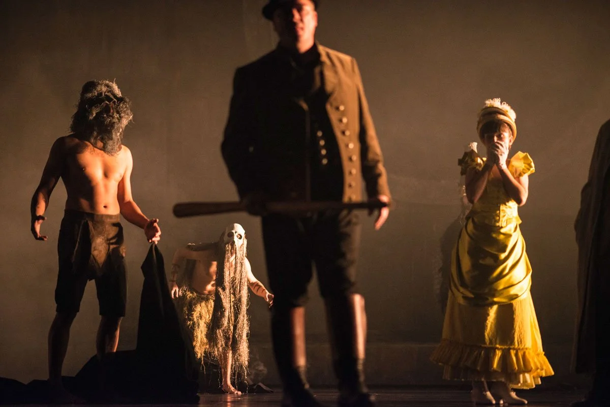 Szenenbild eines Theaterstücks mit vier Figuren in Kostümen, darunter ein Mann mit Zylinder, eine muskulöse Person mit Bart, eine Figur mit grauem, langen Haaren und Maske sowie eine Frau in einem gelben Kleid mit Hand an den Mund, auf dunklem Bühnen