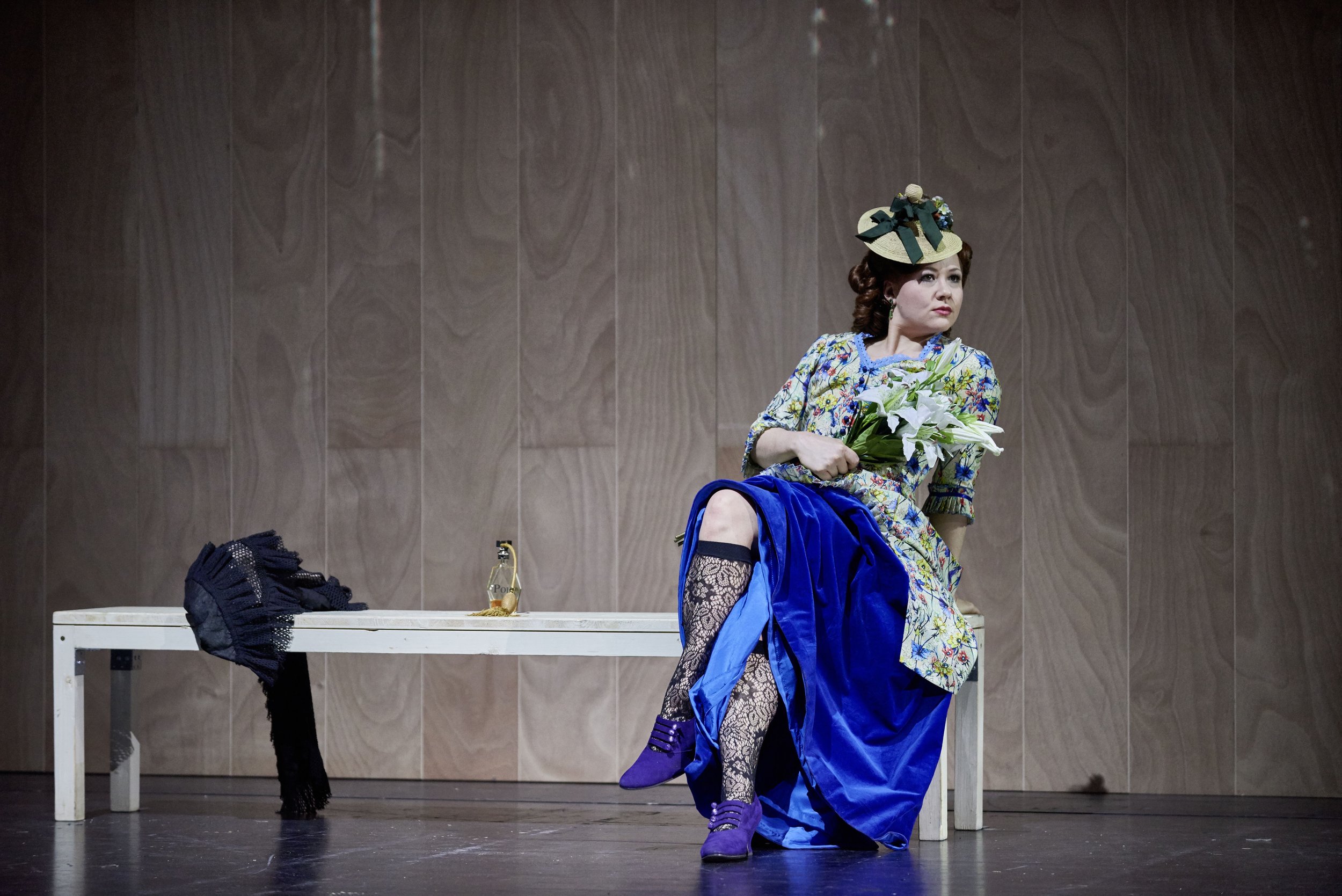 Frau in buntem Kleid mit Hut sitzt auf einer Bank, hält einen Blumenstrauß, trägt schwarze Strümpfe und blaue Schuhe, im Hintergrund eine Holzvertäfelung.