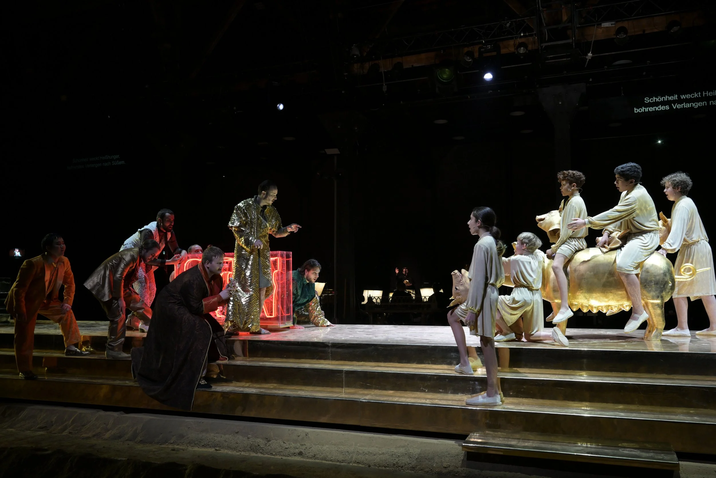 Bühne mit Schauspielern in auffälliger Kleidung, einige sitzen auf einem goldenen Chaiselongue und einige stehen auf Treppen davor, mit dunklem Bühnenhintergrund.