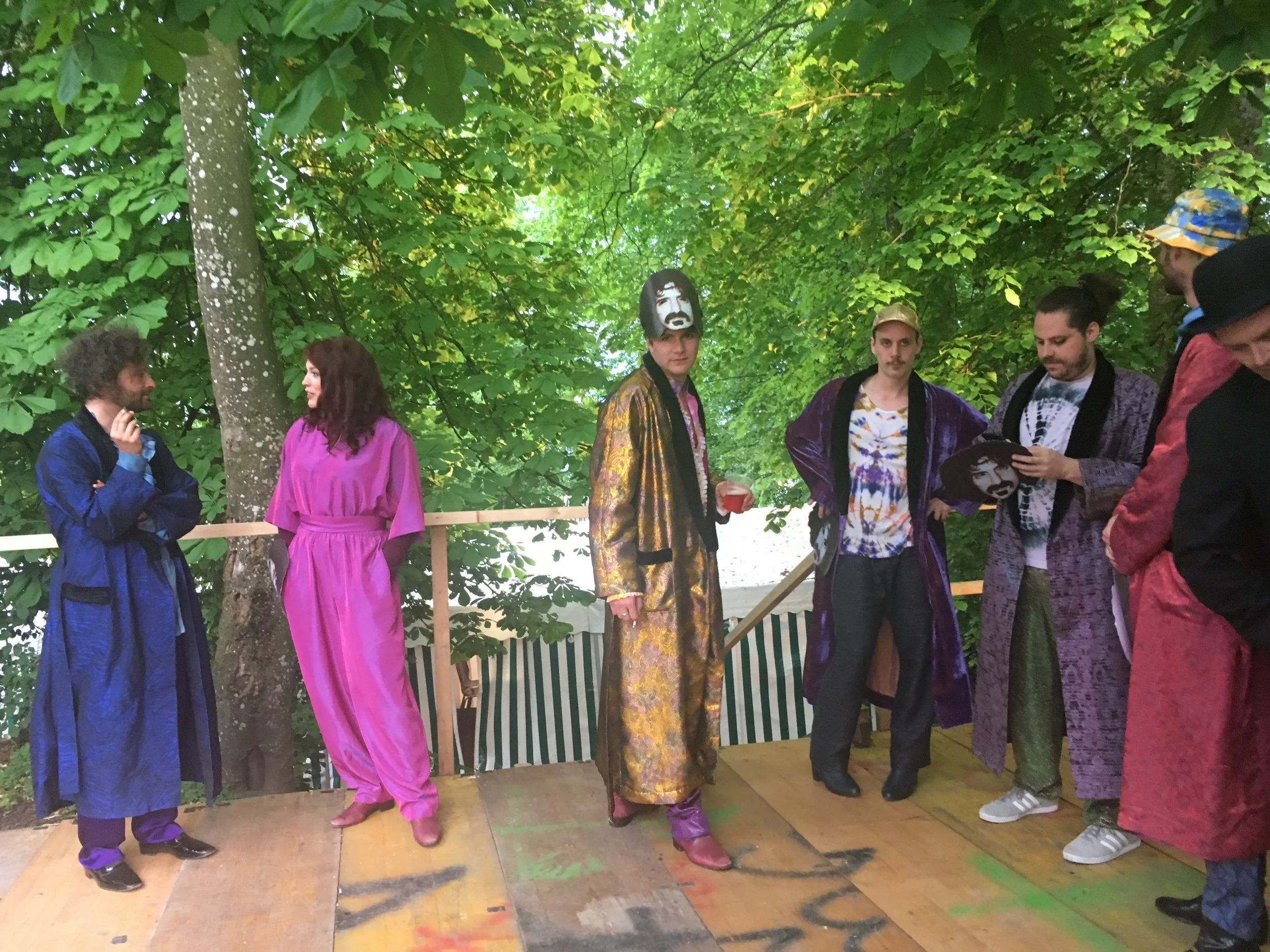 Menschen in bunten, eleganten Kleidern auf einer Holzplattform unter Bäumen