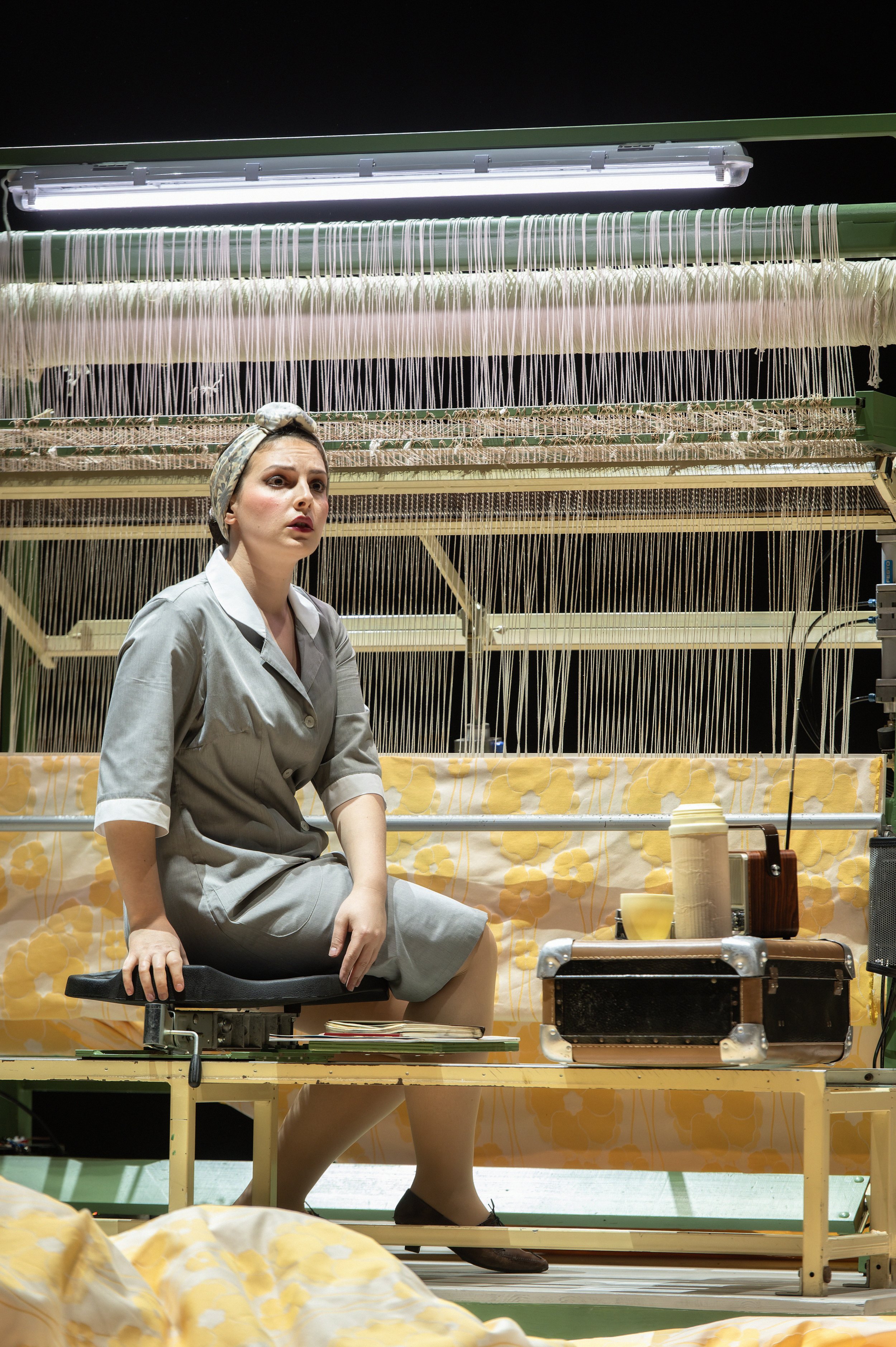 Frau in grauem Kleid sitzt auf einem Stuhl vor einem Nähmaschinen-Tisch, umgeben von Stoffen und Nähutensilien, eine Nähmaschine und Stoffe im Hintergrund.