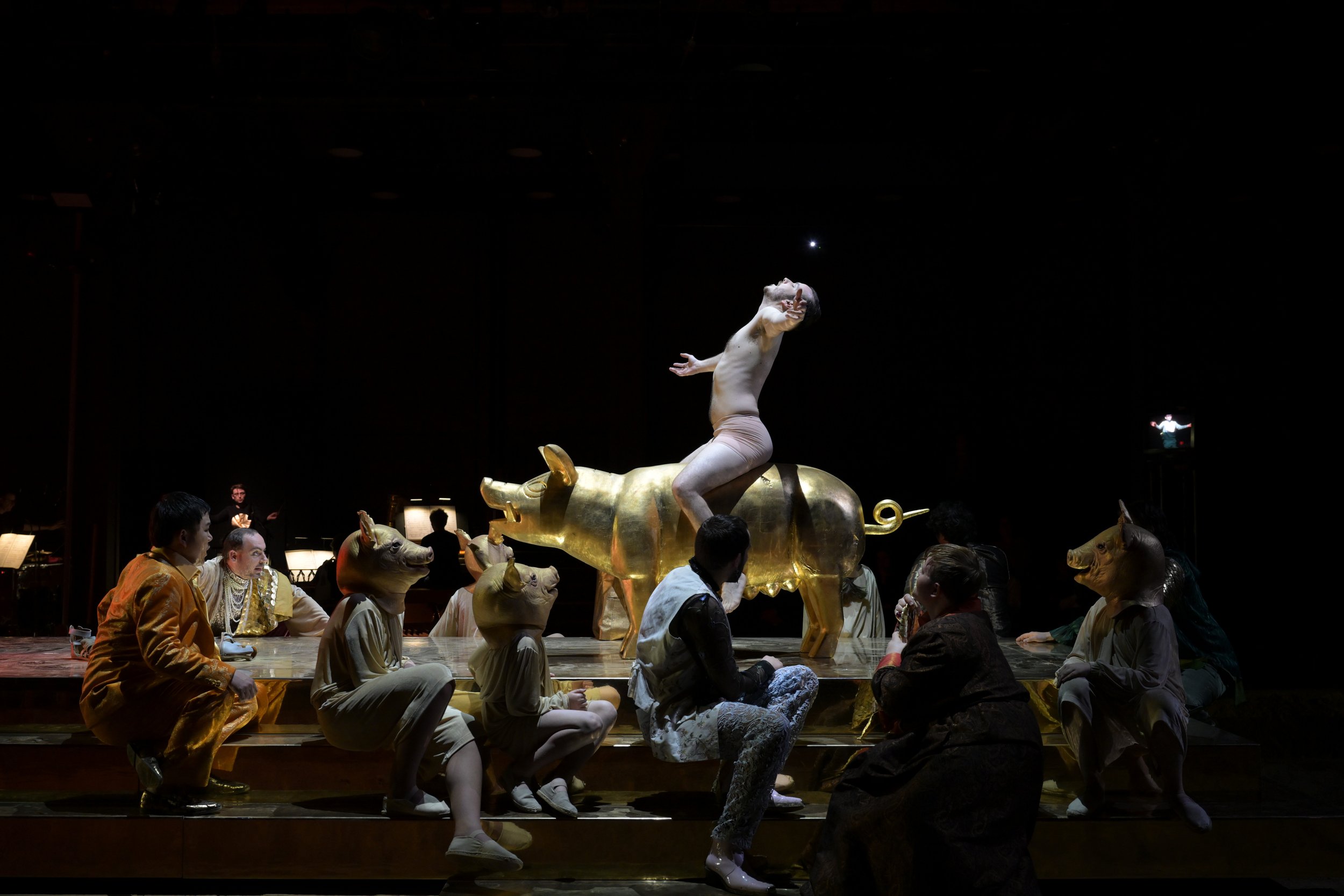 Bühnenaufnahme eines Theaters mit einer Gruppe von Schauspielern in fantastischen Kostümen. Im Mittelpunkt steht eine Figur, die auf einem goldenen Tier sitzt und in die Luft zeigt, während andere Kostüme wie Tiere mit Schwein- und Pferdeköpfen getra