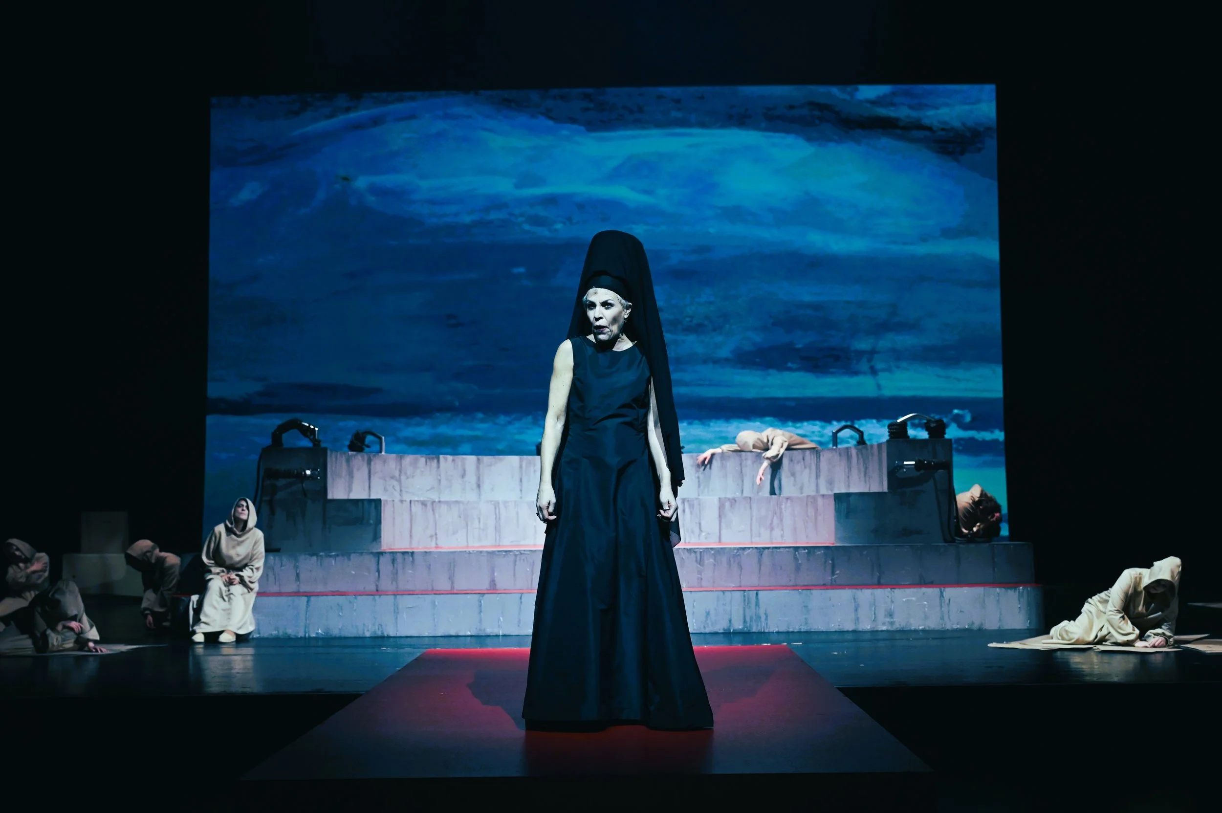 Bühne mit mehreren Schauspielern, eine zentrale Figur in schwarzem Kleid und schwarzen Kopfbedeckung, im Hintergrund ein Meer in einer Kunstprojektion.