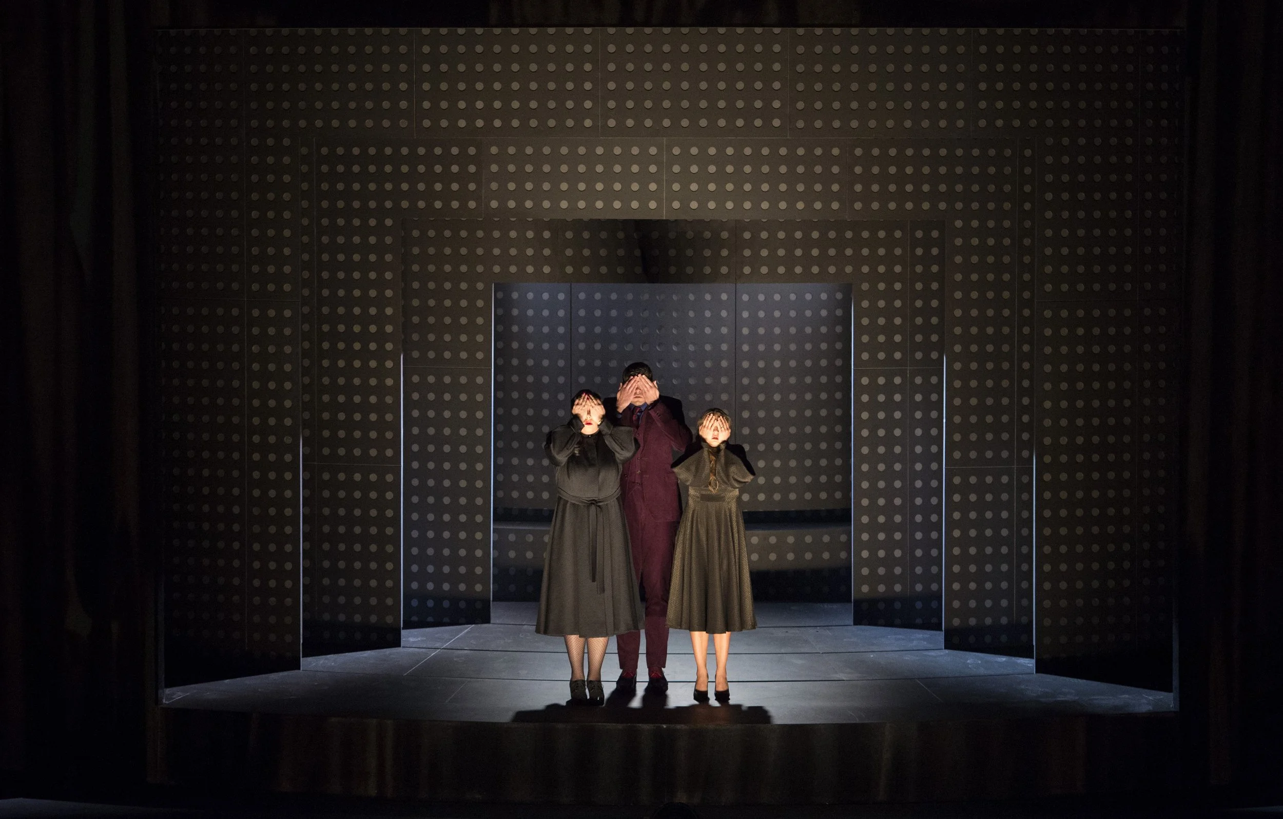Drei Schauspieler stehen auf einer Theaterbühne, die sich hinter schwarzen Kulissen und einer schwarzen Wand mit weißen Punkten befindet, und bedecken ihre Augen.