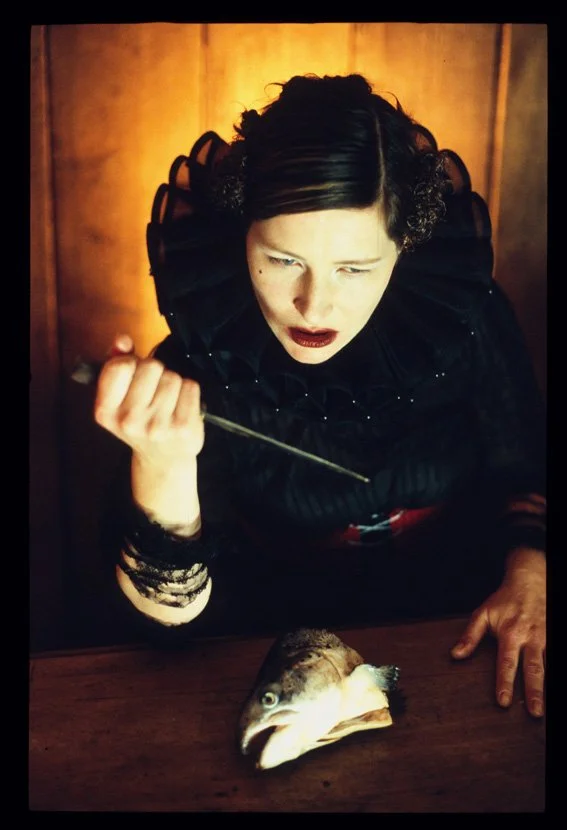 Eine Frau in dunklem viktorianischem Kleid sitzt an einem Tisch mit einem Fischkopf vor ihr, hält eine Dolch in der Hand und schaut angespannt nach oben. Hintergrund aus Holz.