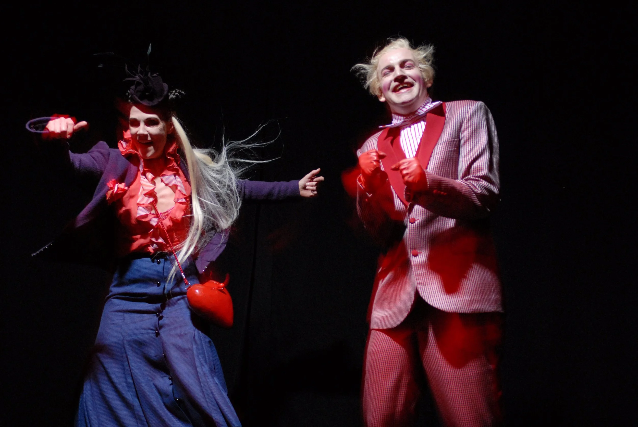 Zwei Schauspieler in farbenfrohen Kostümen auf einer dunklen Bühne, die eine komische Szene darstellen.