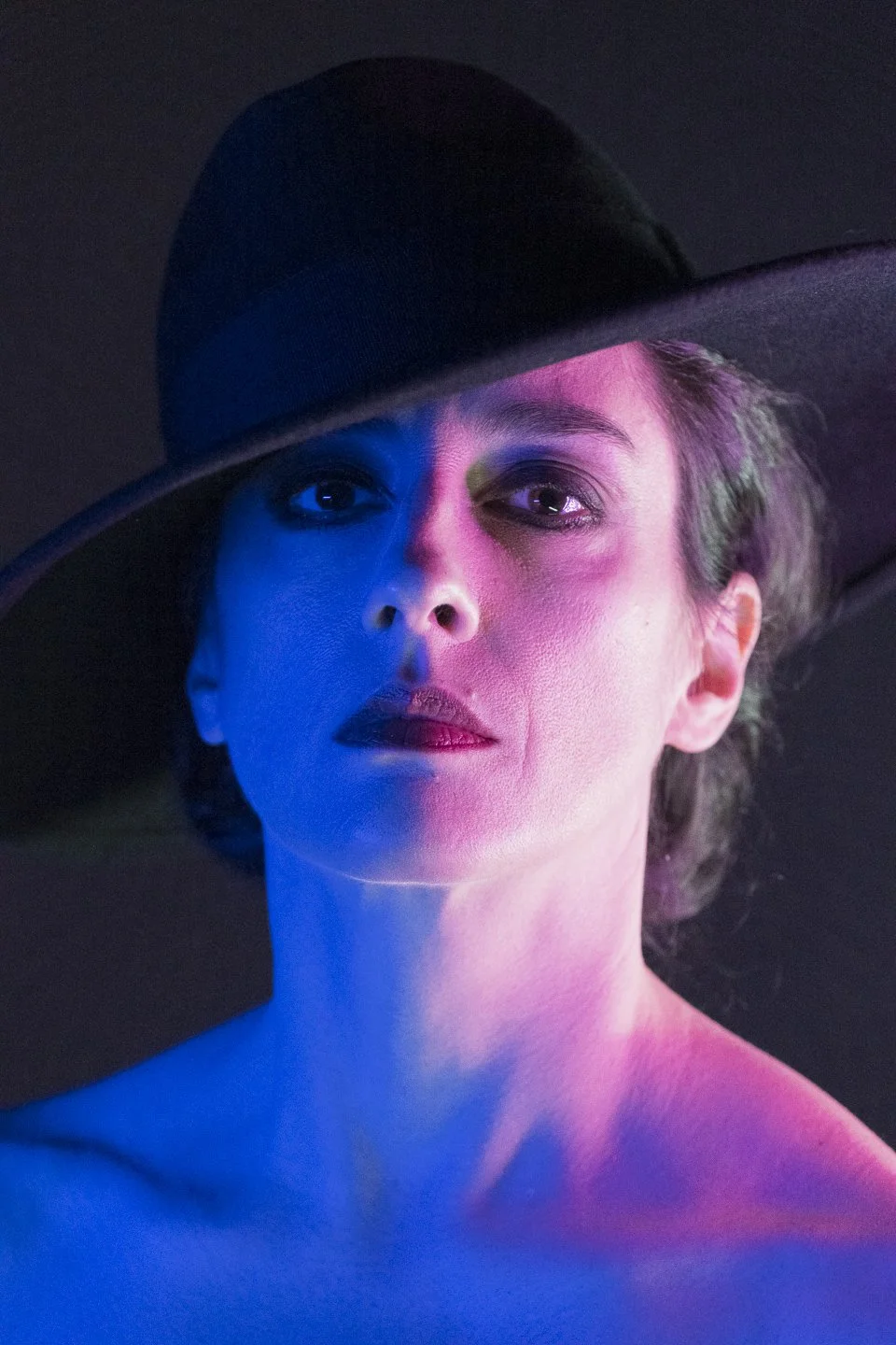 Porträt einer Frau mit eleganten Make-up, halb bedeckt durch einen großen schwarzen Hut, in buntem Licht illuminierte Fotografie.