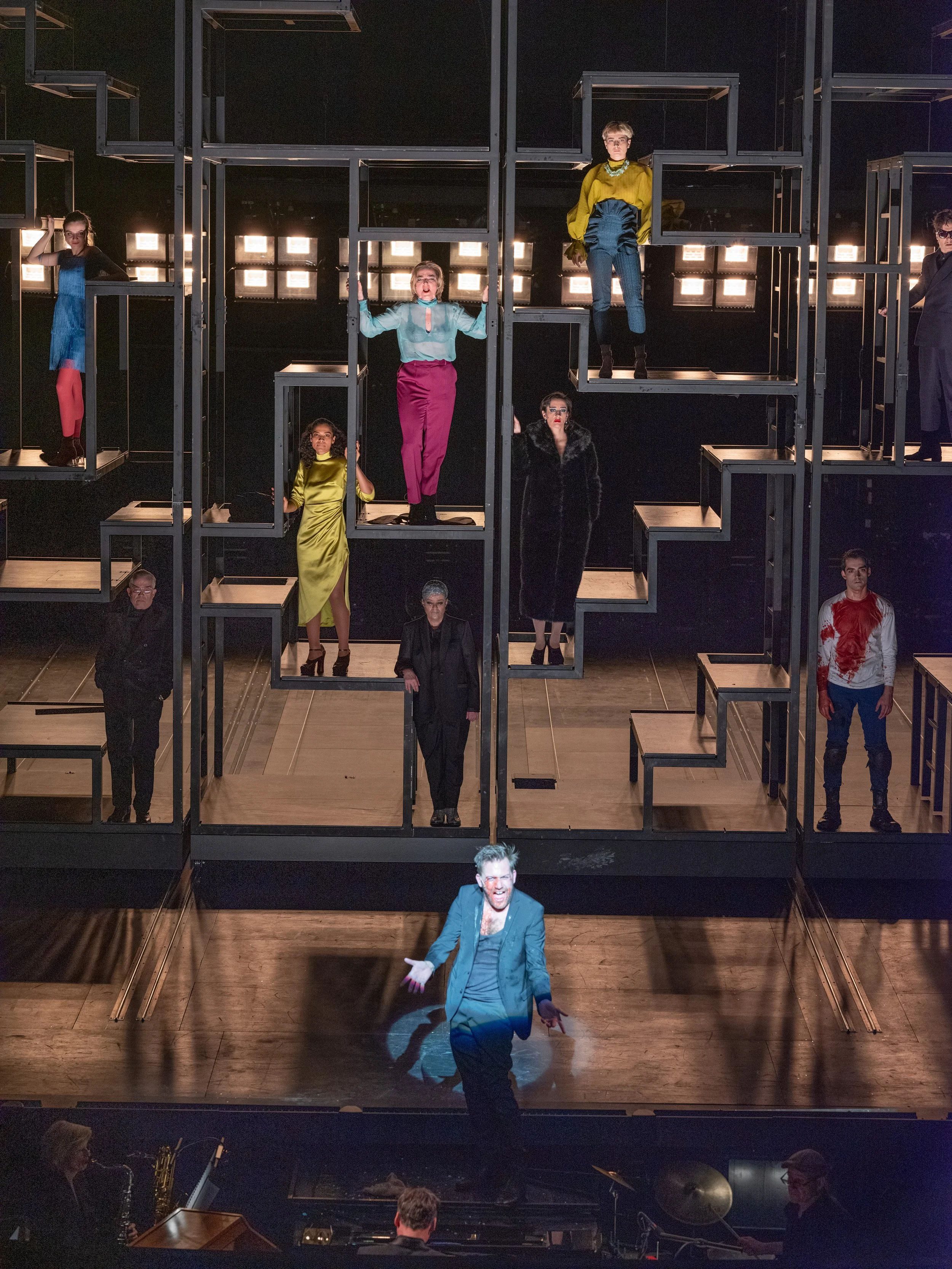 Schauspieler auf einer Bühne, die eine Szene in einem Theaterstück darstellt, mit einer multilevel Kulisse und mehreren Darstellern in bunten Kostümen.