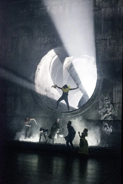 Mehrere Menschen in einer dunklen, industriellen Umgebung, mit einer großen runden Öffnung im Hintergrund und Lichtstrahlen, die durch die Öffnung fallen, während sie tanzen oder sich bewegen.
