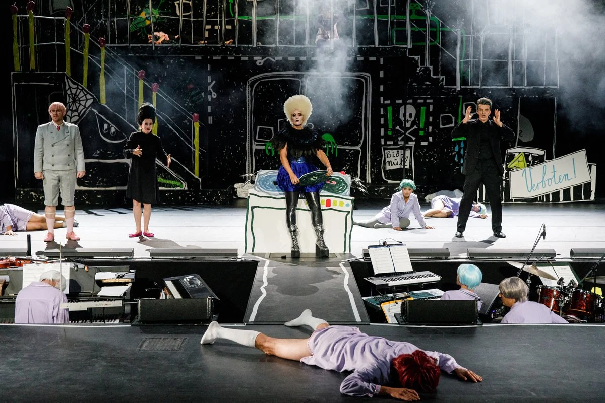 Theaterbühne mit mehreren Darstellern, einer im Mittelpunkt mit Big Hair und schwarzer Kleidung, auf einem Bügel, auf einer kunterbunten Bühne mit Symbolen und Requisiten, im Vordergrund Musiker. Scheinwerfer und Nebel verstärken den Bühnenmoment.