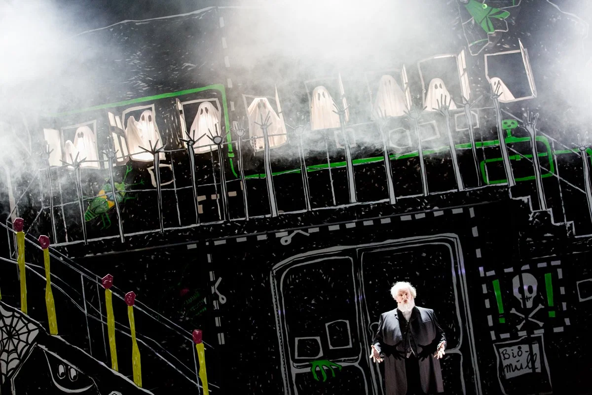Ein Bühnenszene mit einer Person in schwarzer Kleidung vor einer schwarz-weißen Kulisse, die grüne und schwarze Details aufweist. Es gibt Puppen in Geisterkostümen und Spinnennetze, die an eine Halloween- oder Geisterthematik erinnern.