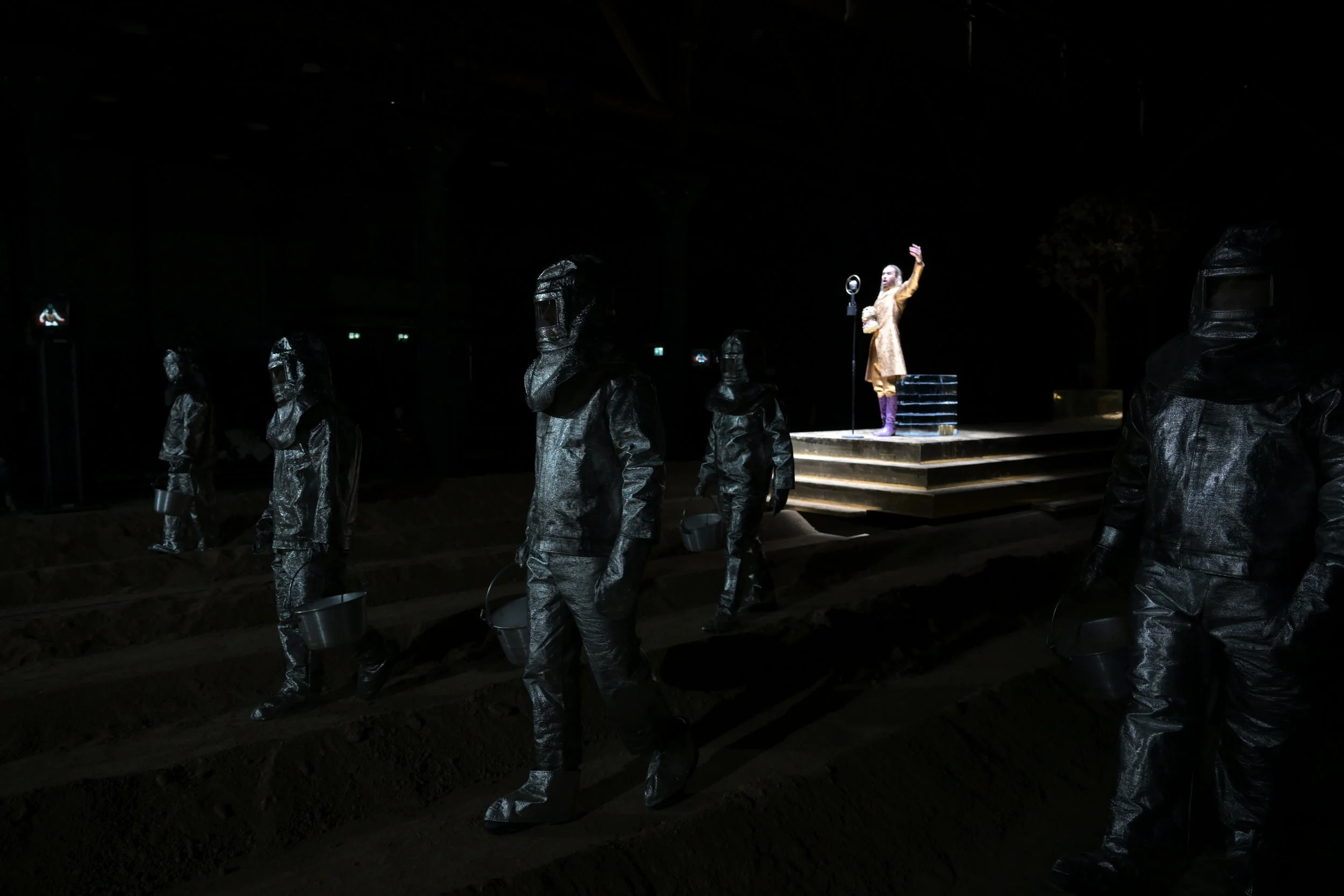 Menschen in schwarzen Schutzanzügen stehen auf einer dunklen Bühne, im Hintergrund eine Person auf einem erhöhten Podest, die eine Hand hebt.