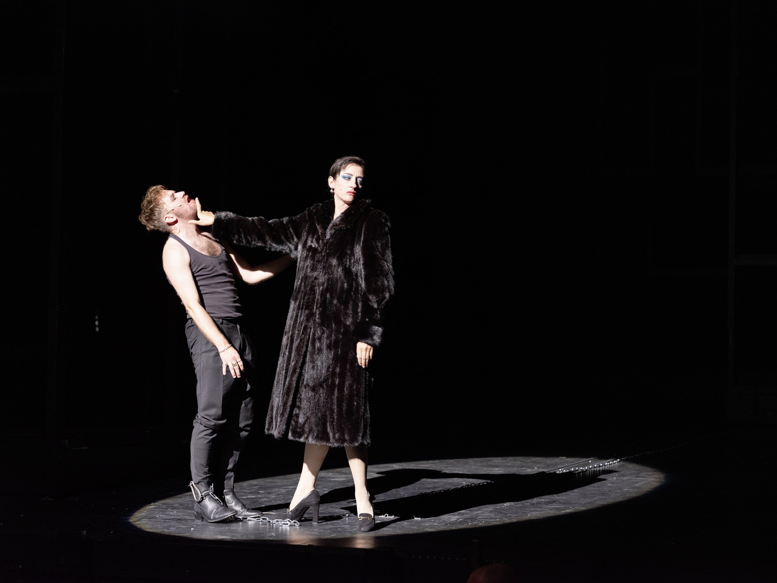 Zwei Menschen in einer dramatischen Szene auf einer dunklen Bühne. Ein Mann trägt eine schwarze Tanktop und schwarze Hosen, während die andere Person in einem langen schwarzen Mantel steht und eine dominante Haltung zeigt.