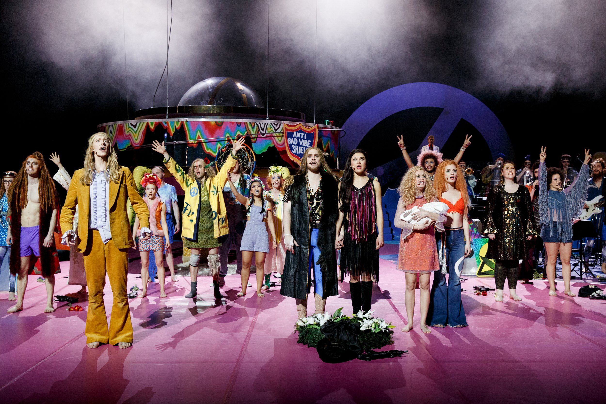 Schauspieler auf der Bühne bei einer Theaterproduktion, umgeben von anderen Darstellern, im Hintergrund ein farbenfrohes Bühnenbild mit einem Schild »Anti Bad Vibe Show«. Der Boden ist pink, und die Atmosphäre wirkt dramatisch und lebhaft.