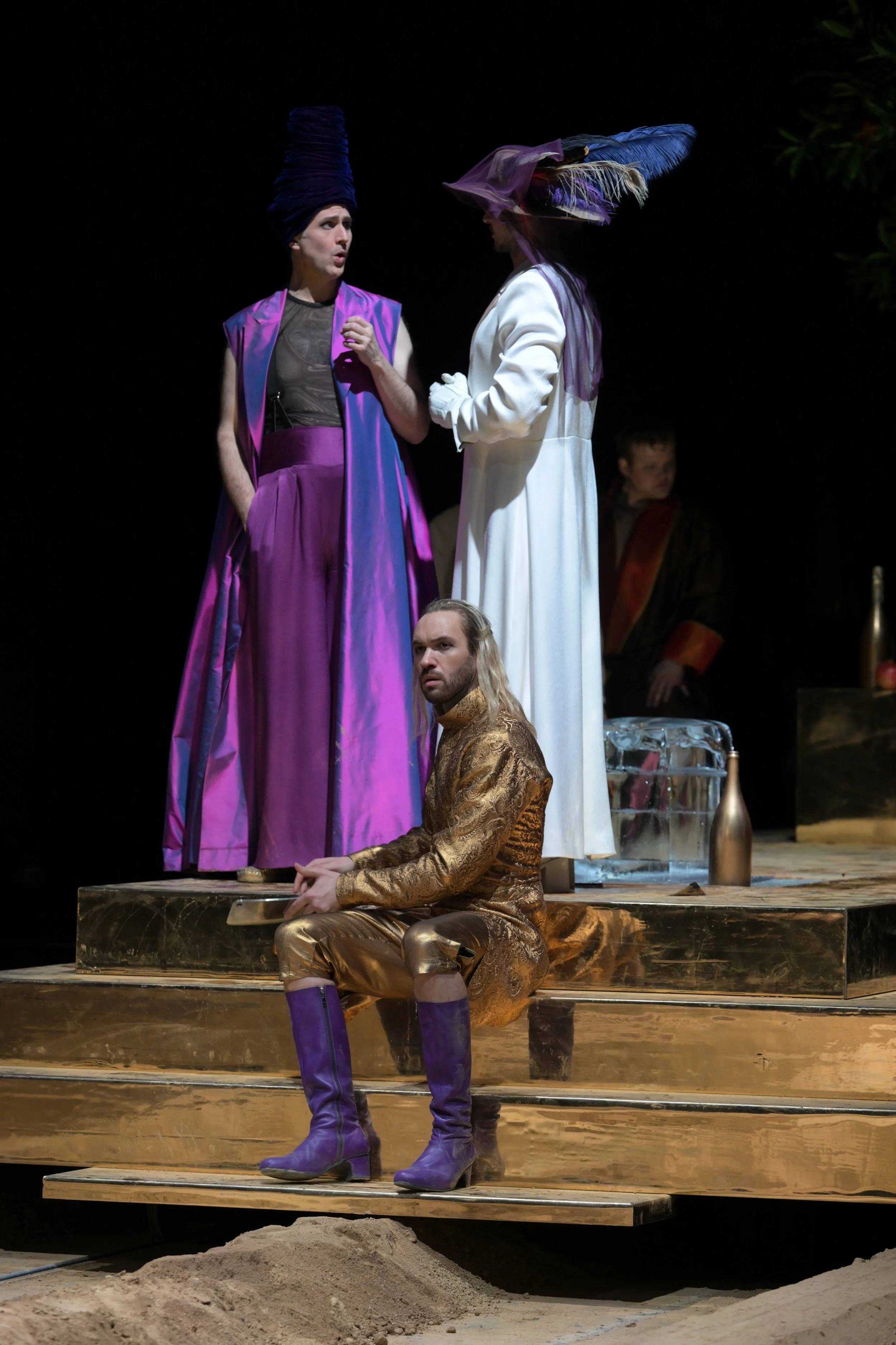 Szenenaufnahme aus einem Theaterstück mit vier Schauspielern in extravaganten Kostümen, die auf einer Bühne mit goldfarbenem Boden und Sand im Vordergrund agieren. Zwei Schauspieler stehen auf einem erhöhten Teil der Bühne, einer sitzt auf der Stufe,