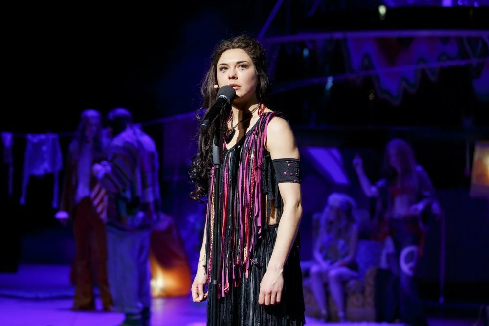 Junge Frau singt bei einer Theateraufführung, im Hintergrund sind andere Darsteller und Bühnenrequisiten zu sehen.