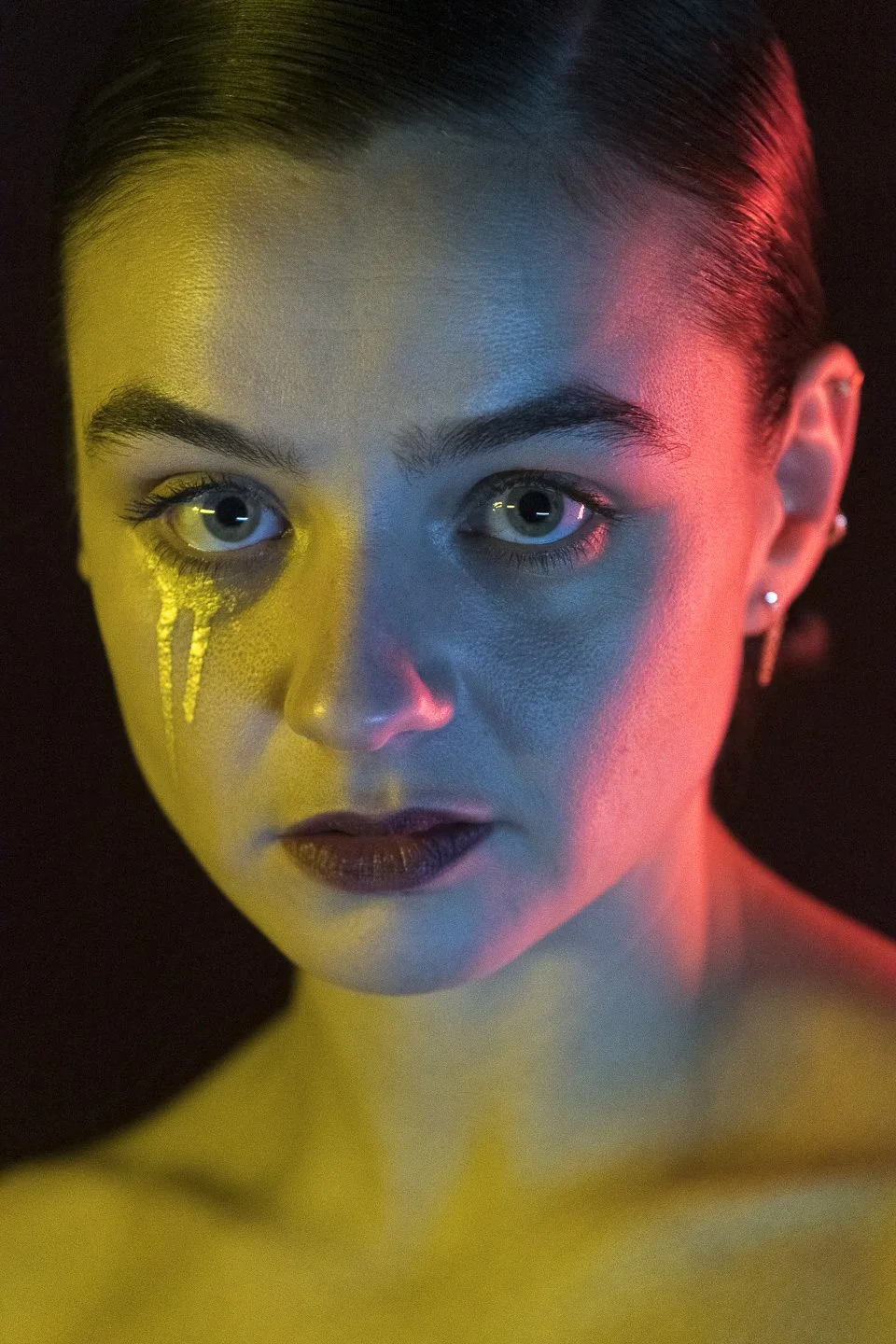 Nahaufnahme eines jungen Frauengesichts mit Make-up, silbernen Ohrringen und Neonlichtern in Blau, Gelb und Rot