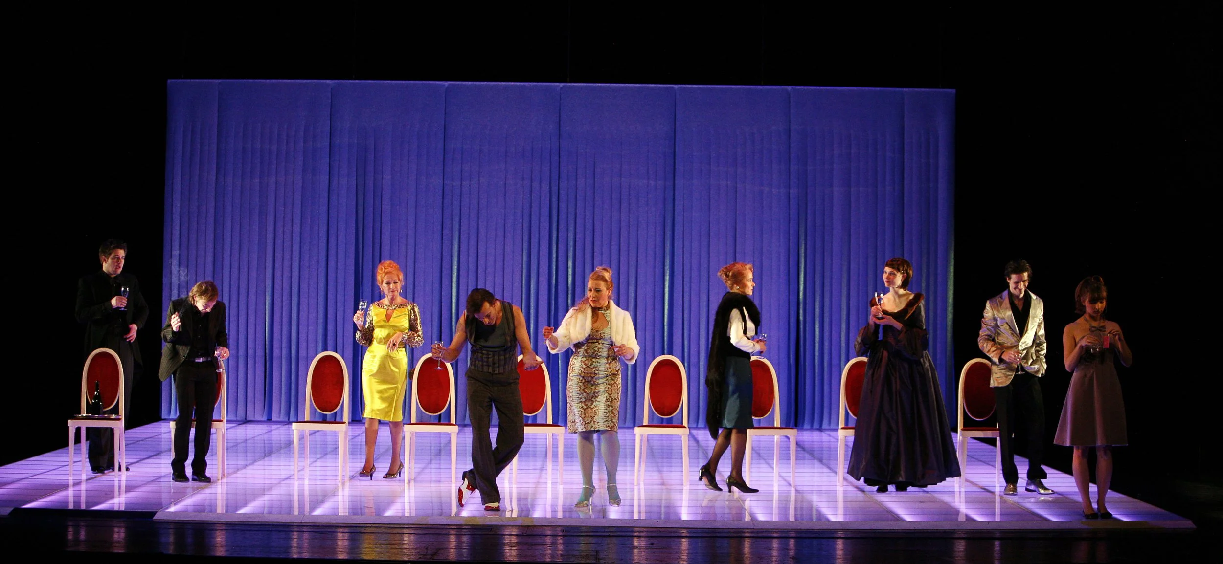 Bühnenaufführung mit neun Schauspielern, die auf einer Bühne mit blauer Kulisse stehen, einige halten Gläser mit Getränken, Bühnenbeleuchtung und leeres Stuhl im Vordergrund.