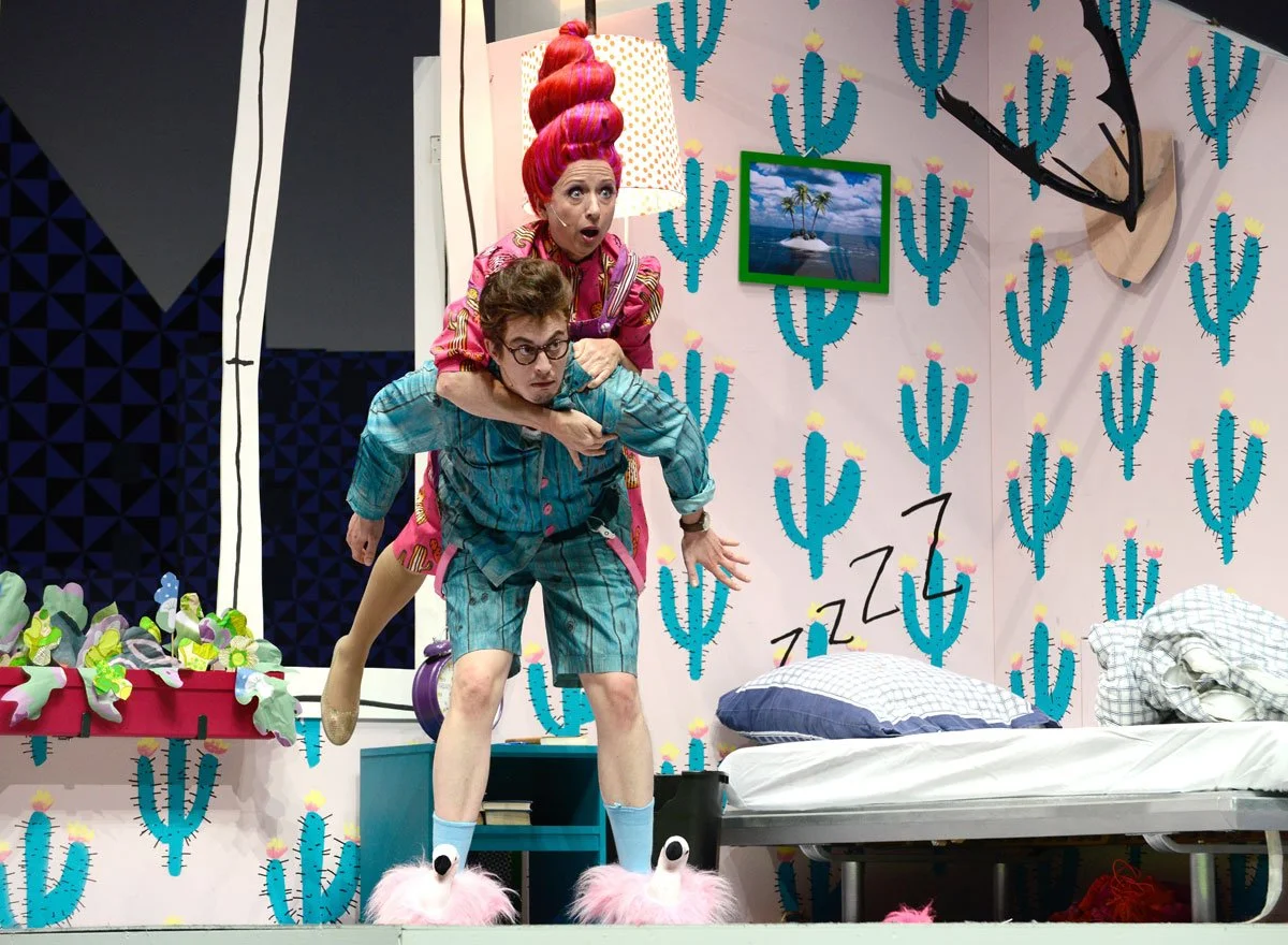 zwei Schauspieler in einem surrealen Schlafzimmer. Die Frau trägt große, pinke, gewellte Frisur und pinkes Kleid, die Männer trägt eine blaue Schlafkleidung und pinke Flamingo-Schlappen. Es gibt eine Wand mit Kaktus-Motiv, eine Bett mit Kissen, eine 