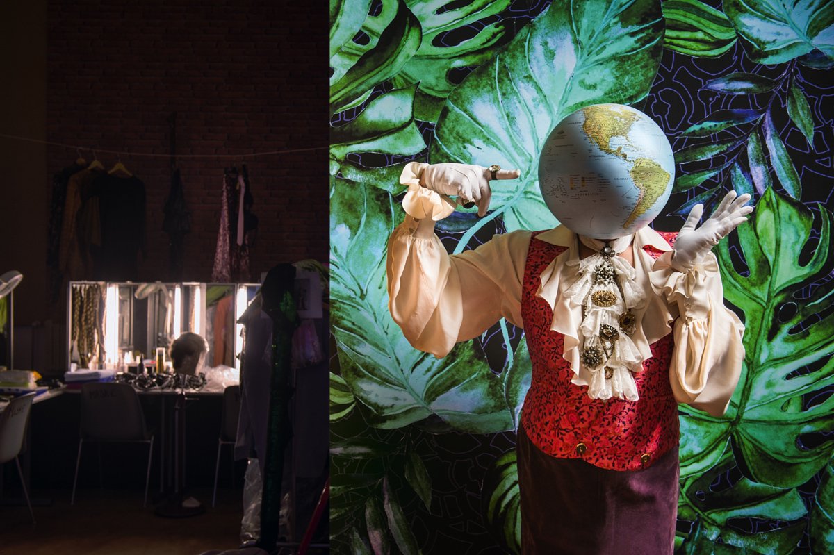 Person mit Globus im Gesicht, gekleidet im historischen Kostüm, vor einer Wand mit großen grünen Blättern im Hintergrund.