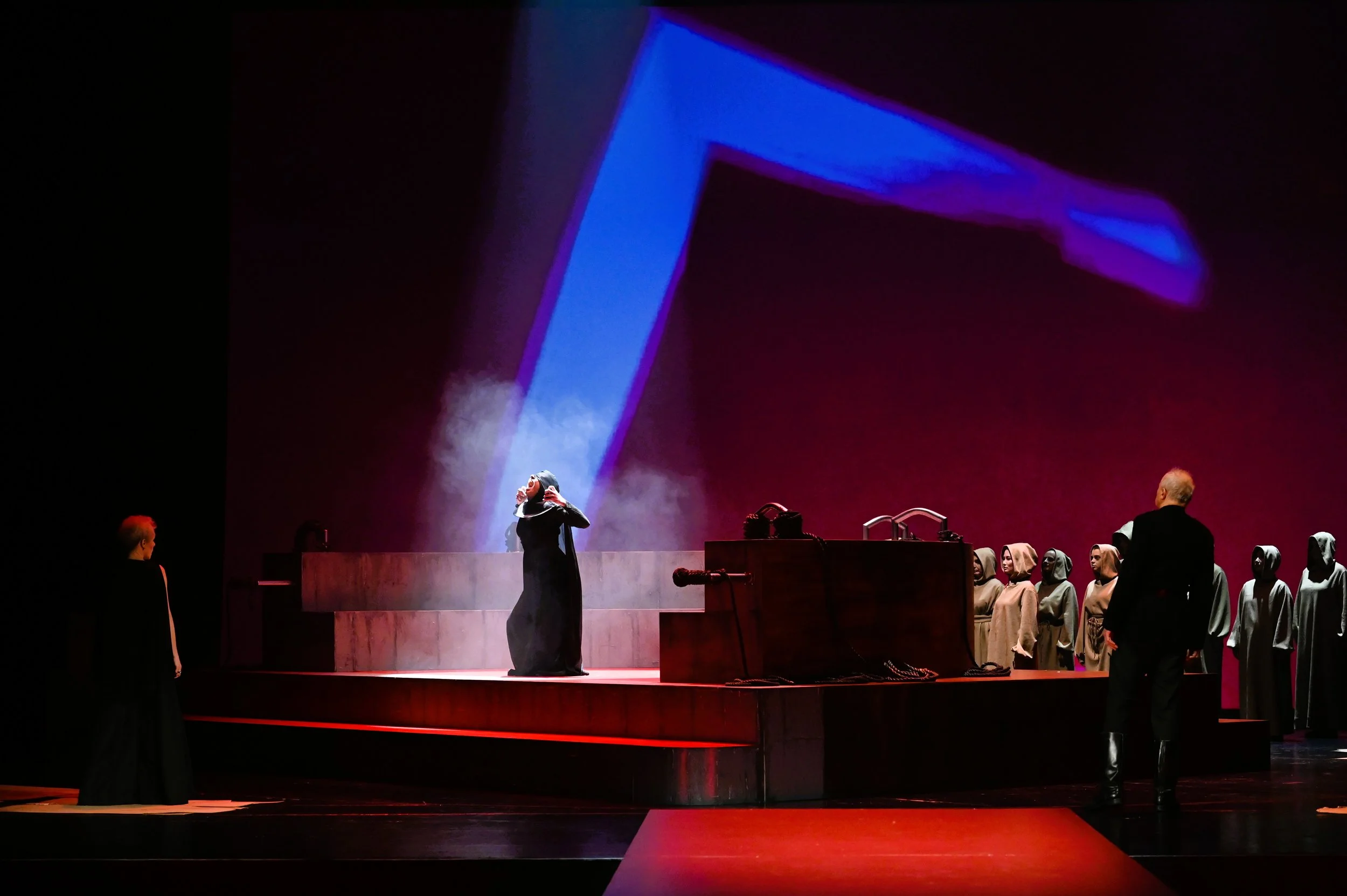 Eine Theaterszene mit mehreren Darstellern, die auf einer Bühne stehen. Im Hintergrund ist eine große, leuchtende Projektion eines blauen Gegenstands zu sehen. Die Szene ist dunkel, das Licht fokussiert auf die Akteure.