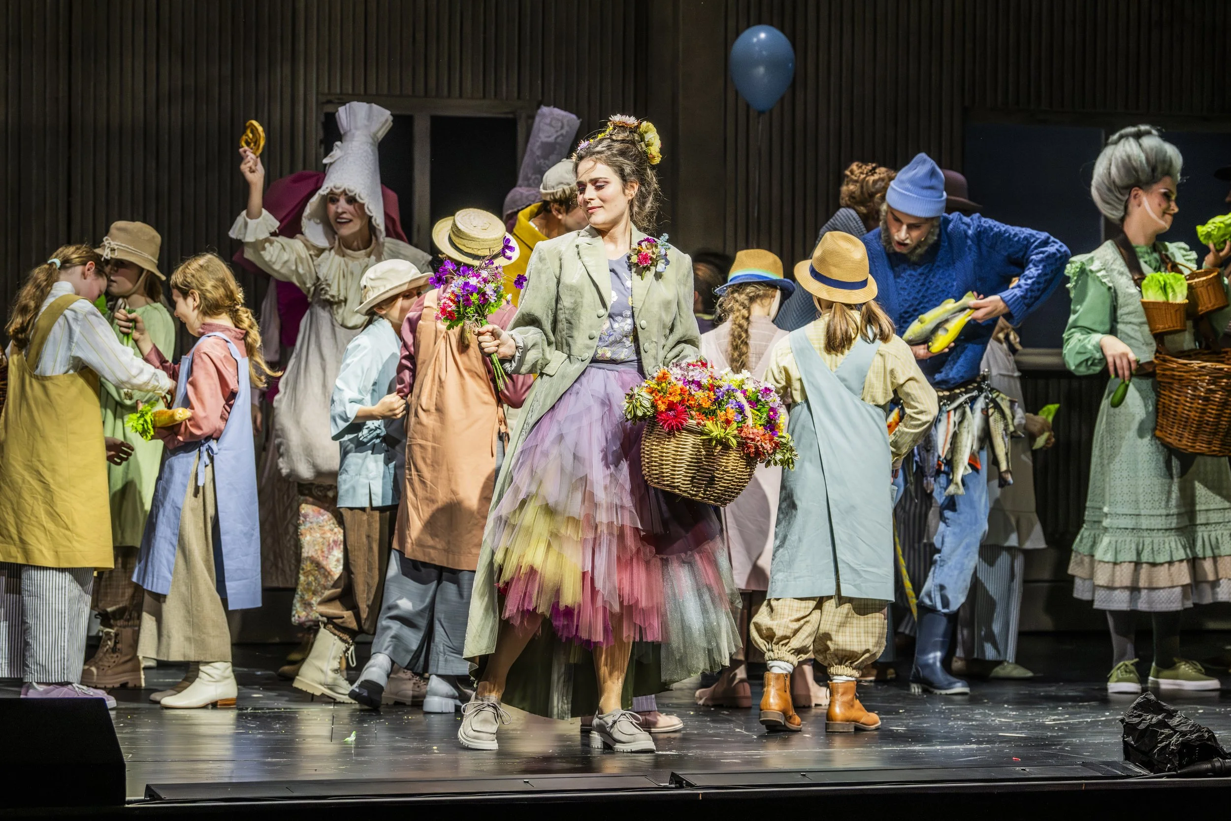 Theaterstück mit mehreren schauspielern in bunten Kostümen auf Bühne, zentrale Frau mit Blumen und Korb im Vordergrund.