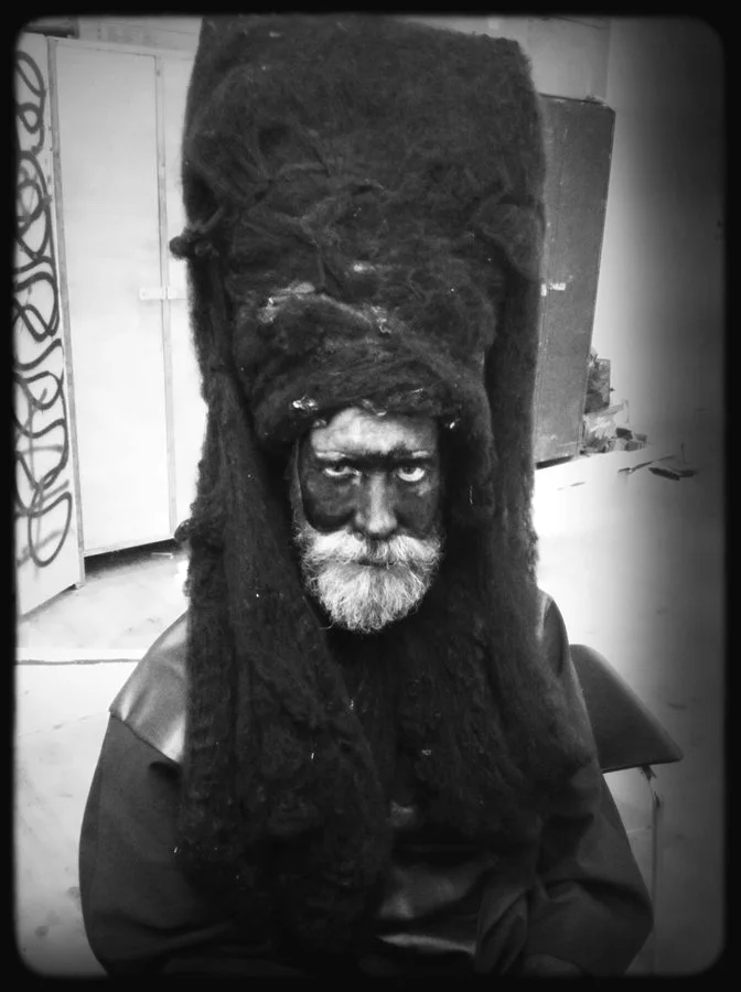 Schwarz-weiß-Fotografie eines Mannes mit grauem Bart und dunklem Gesicht, der ein kunstvoll gestaltetes, großes Haarschmuckstück trägt, das an ein Kopfstück erinnert.