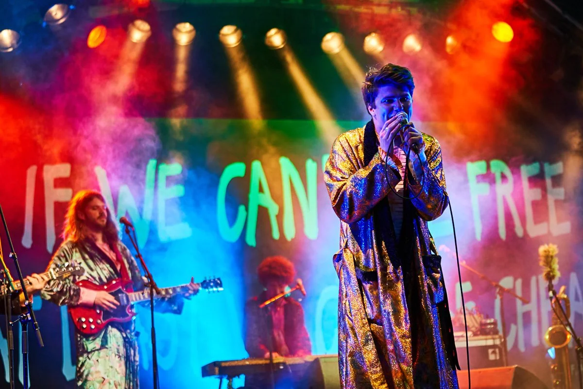 Auf einer Bühne spielen Musiker mit bunten Lichteffekten. Im Vordergrund singt ein Mann mit dunklen Haaren und goldenem, glänzendem Mantel. Im Hintergrund sind weitere Musiker, einer mit Gitarrenmusikinstrumenten und ein weiterer an einem Keyboard. E