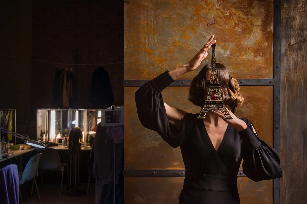 Frau mit modellierter Eiffelturm-Statue vor dem Gesicht in einem Atelier mit Schminktischen im Hintergrund.