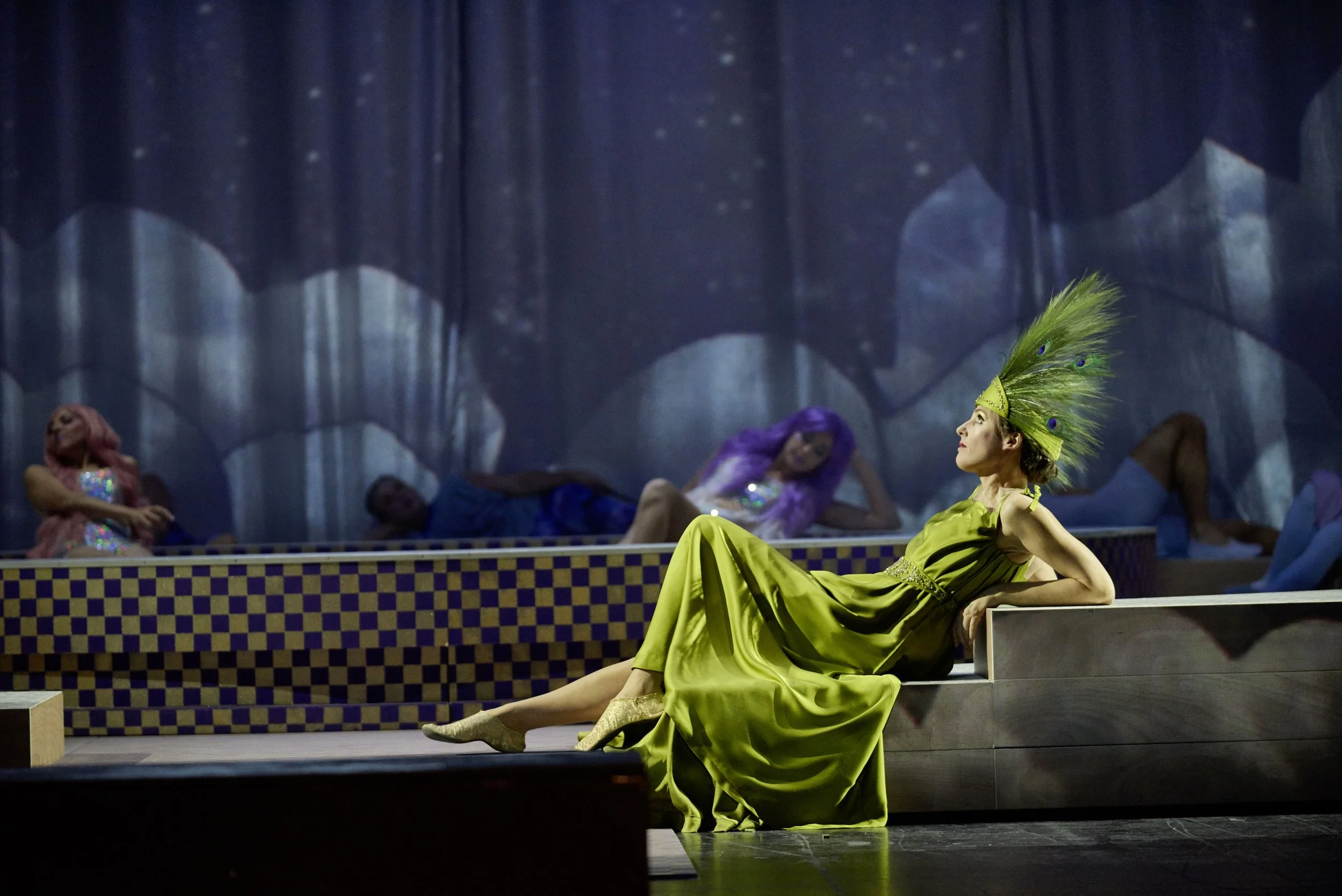 Eine Frau in einem grünen Kleid mit einem aufwändigen Federschmuck sitzt entspannt auf einer Bühne, im Hintergrund liegen und sitzen andere Frauen mit bunten Haaren und glitzernden Kleidern in einer künstlerischen Szene.