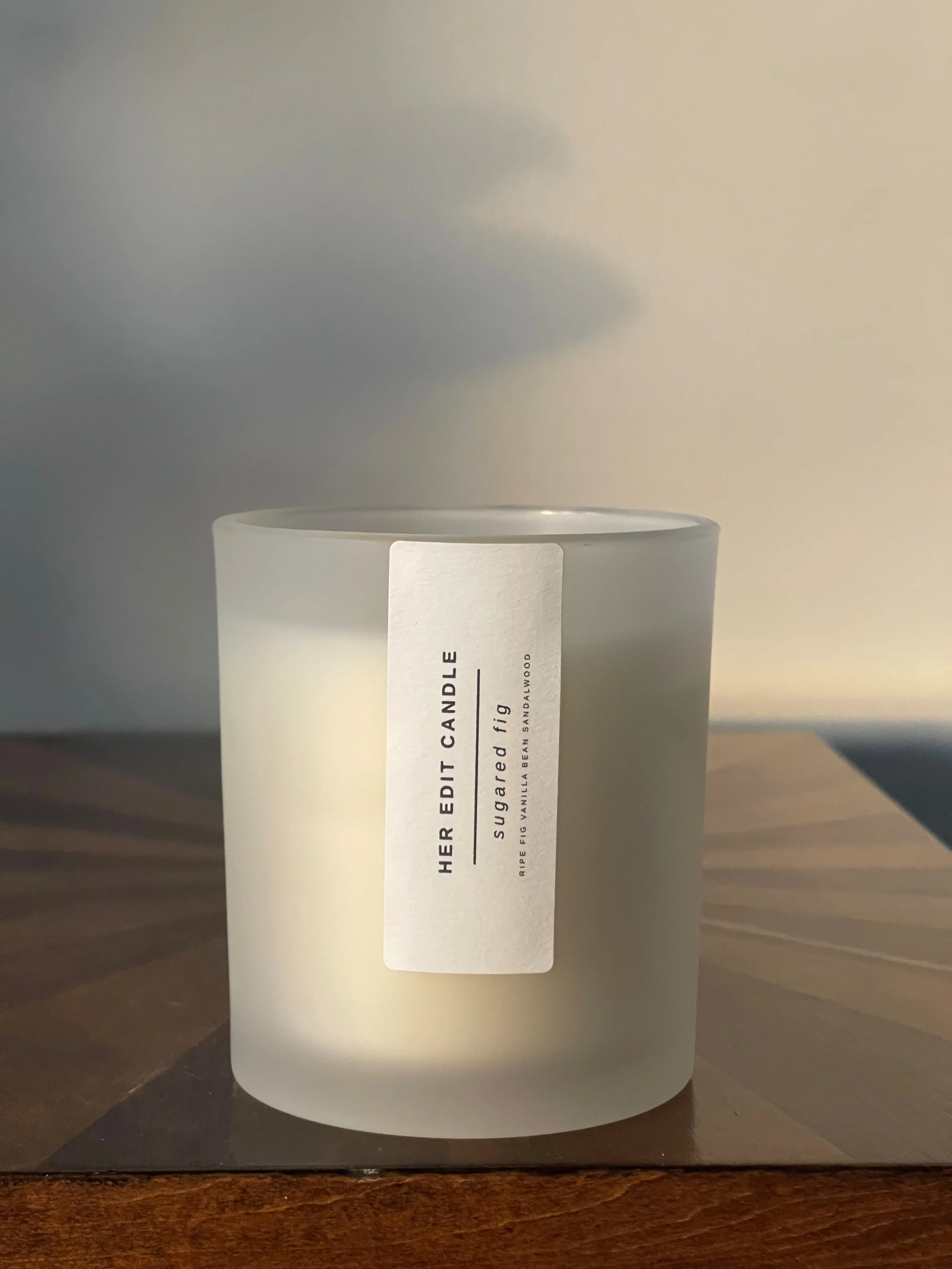 A white frosted glass candle with a label that reads HER EDIT CANDLE, scented with sugared fig, with notes of ripe fig, vanilla bean, sandalwood, placed on a wooden surface.